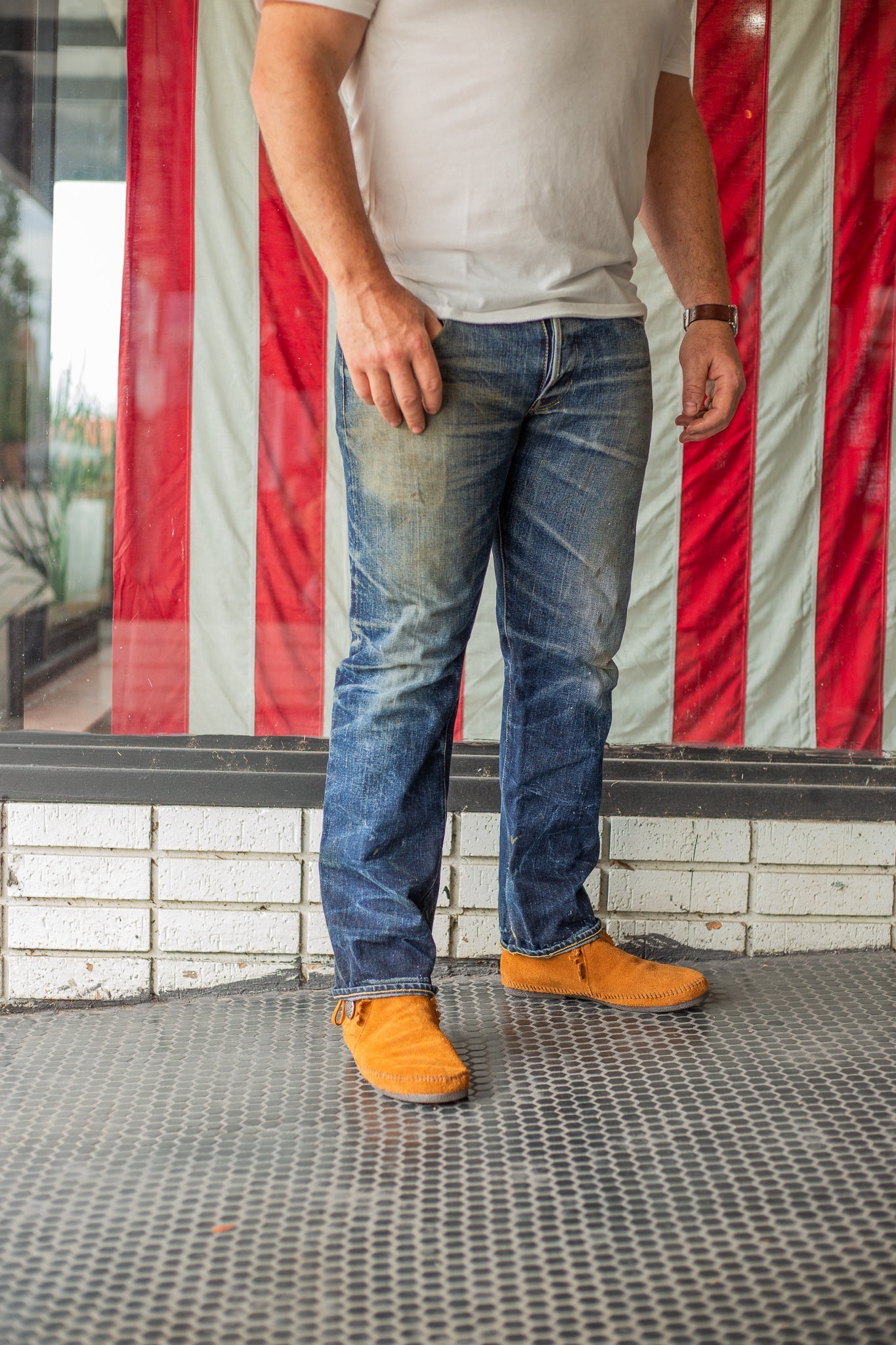 Man wearing white t-shirt, worn blue jeans, and orange moccasin shoes standing on patterned tile floor