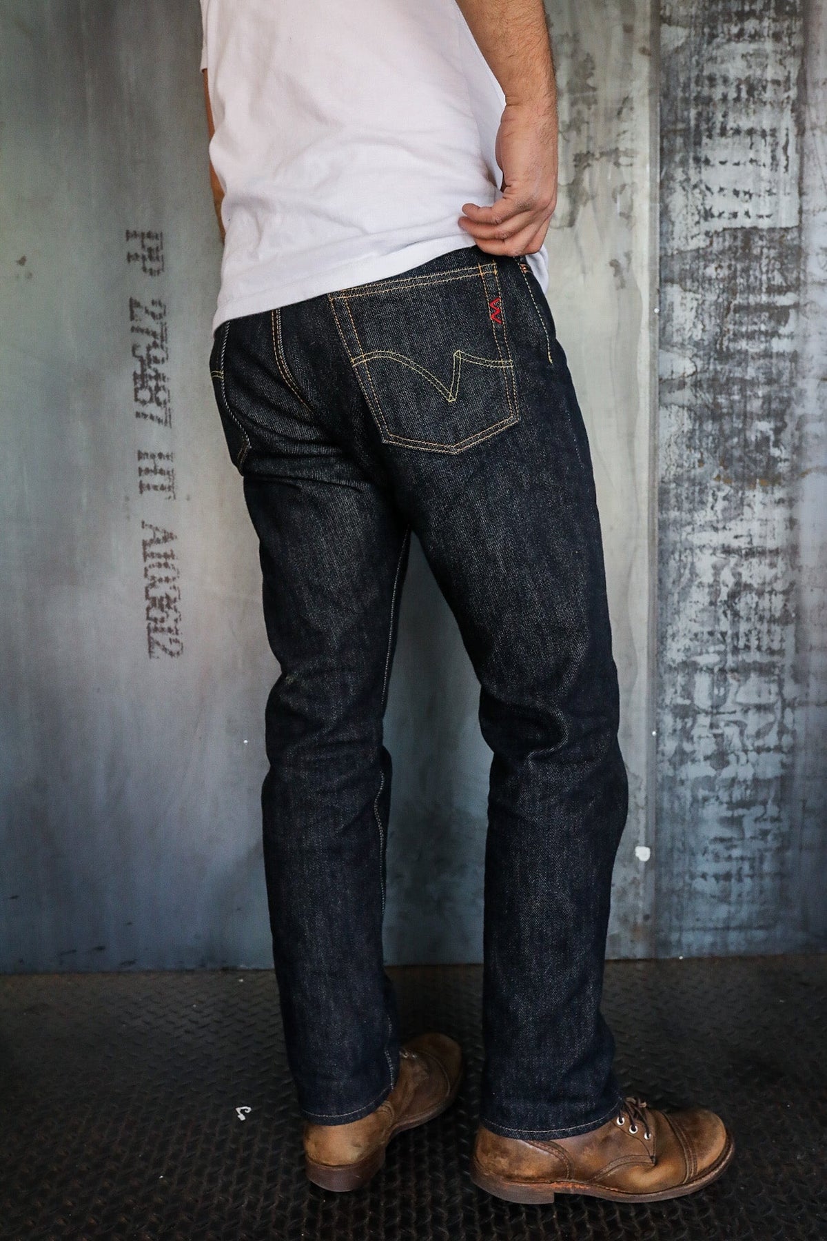 Man wearing dark raw denim jeans and brown leather boots standing on metal floor with industrial gray background