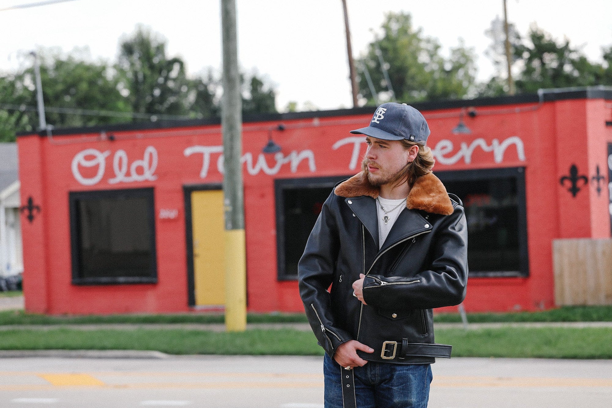 Man wearing black leather jacket with brown fur collar and blue cap standing in front of red building with Old Town Tavern sign