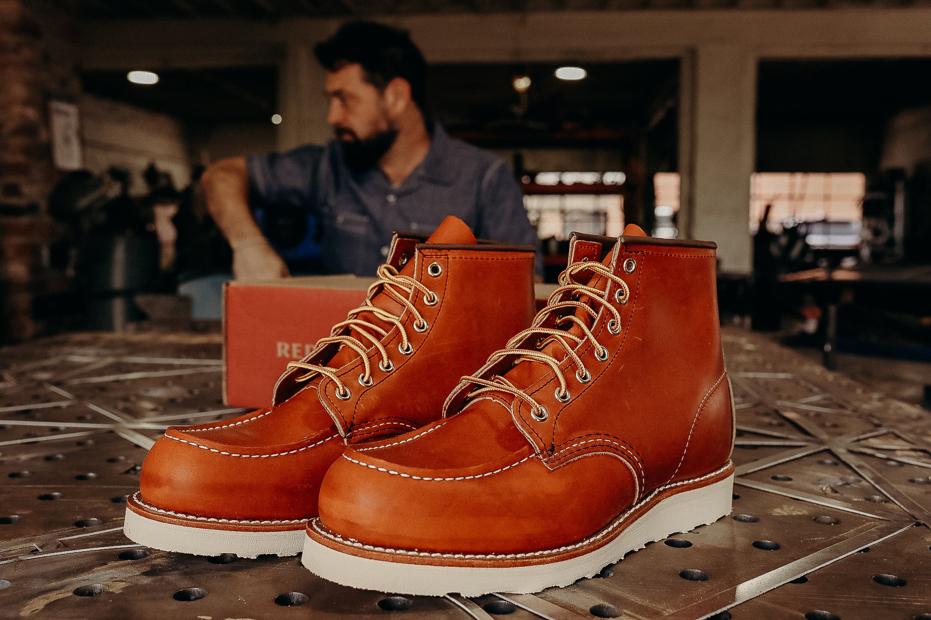 Pair of rugged brown leather work boots with white soles on metal table and man in workshop background