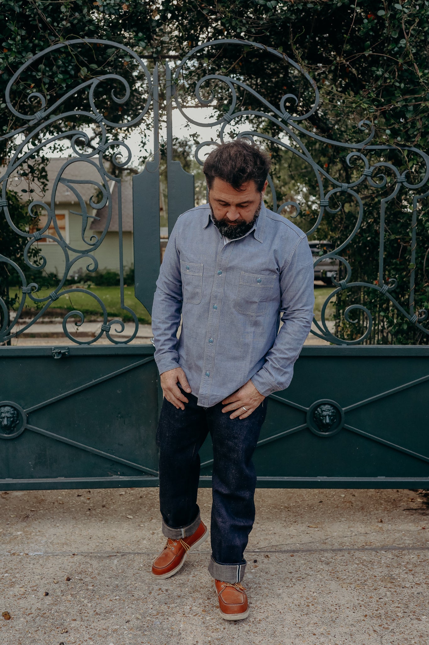 Man in casual denim shirt, dark jeans, and brown leather boots standing in front of decorative wrought iron gate