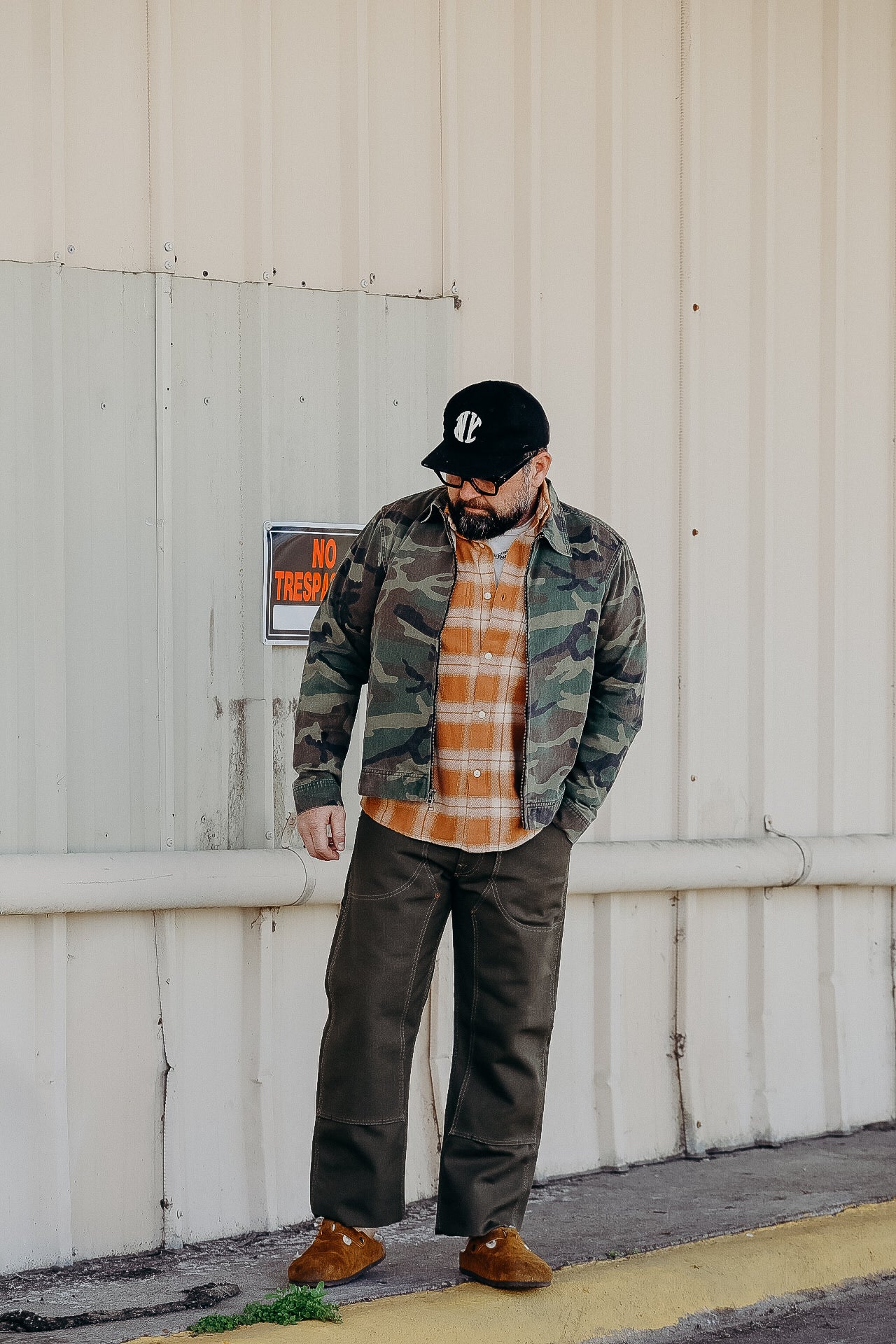 Man wearing camo jacket, orange plaid shirt, work pants, brown shoes, and black cap standing by industrial wall with no trespassing sign