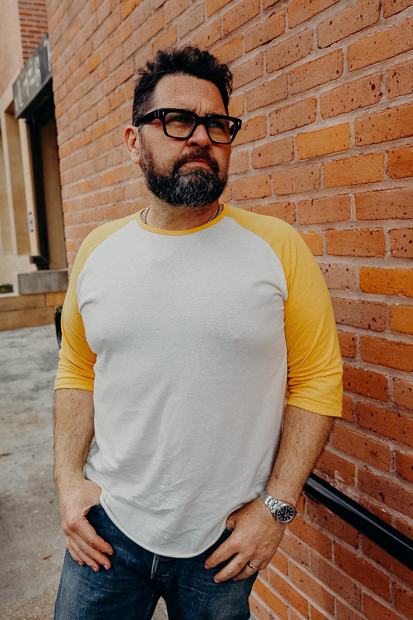 Man with beard and glasses wearing yellow and white raglan shirt, standing against brick wall outdoors
