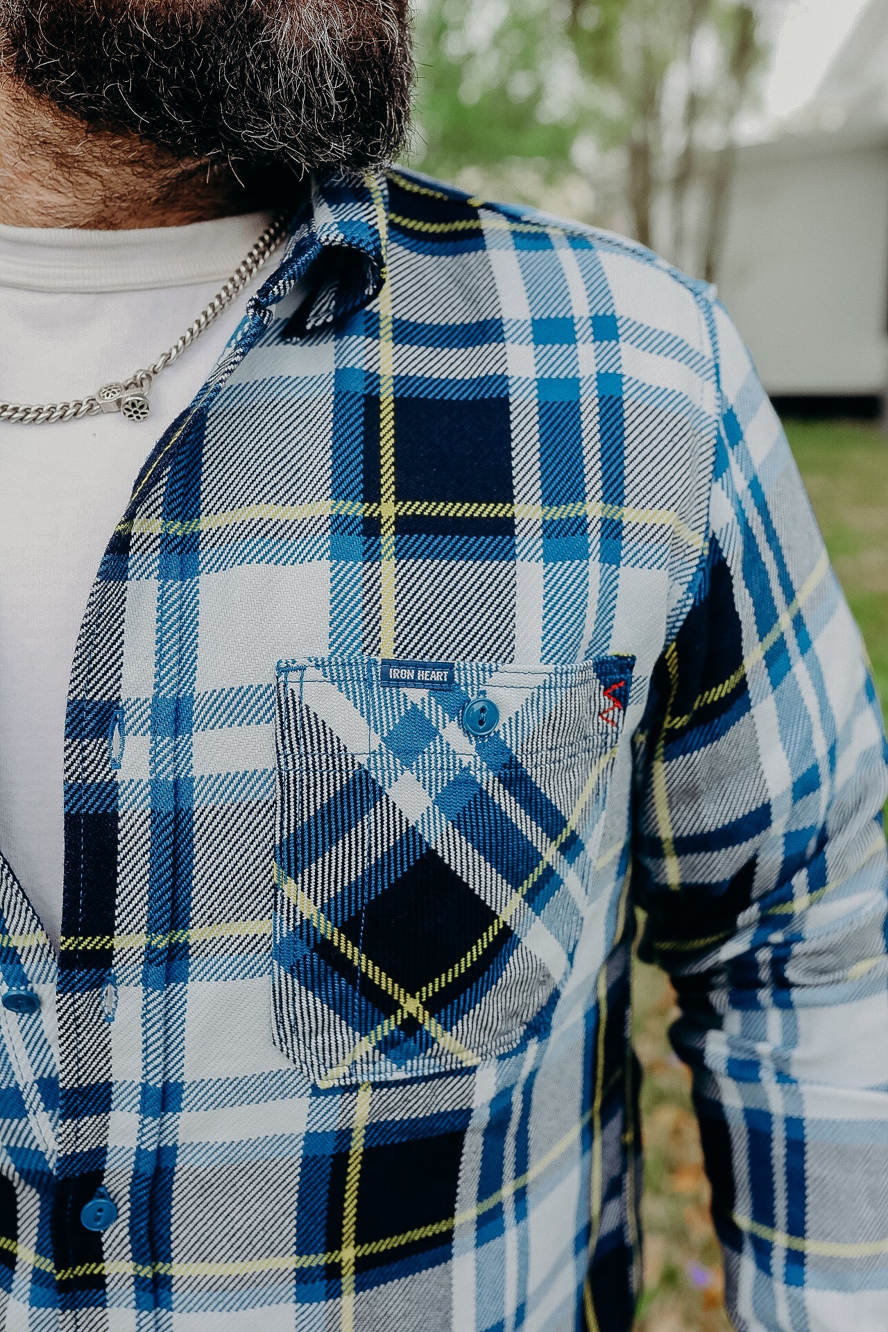 Close-up of a man wearing a blue, white, and yellow plaid Iron Heart shirt with a chain necklace outdoors