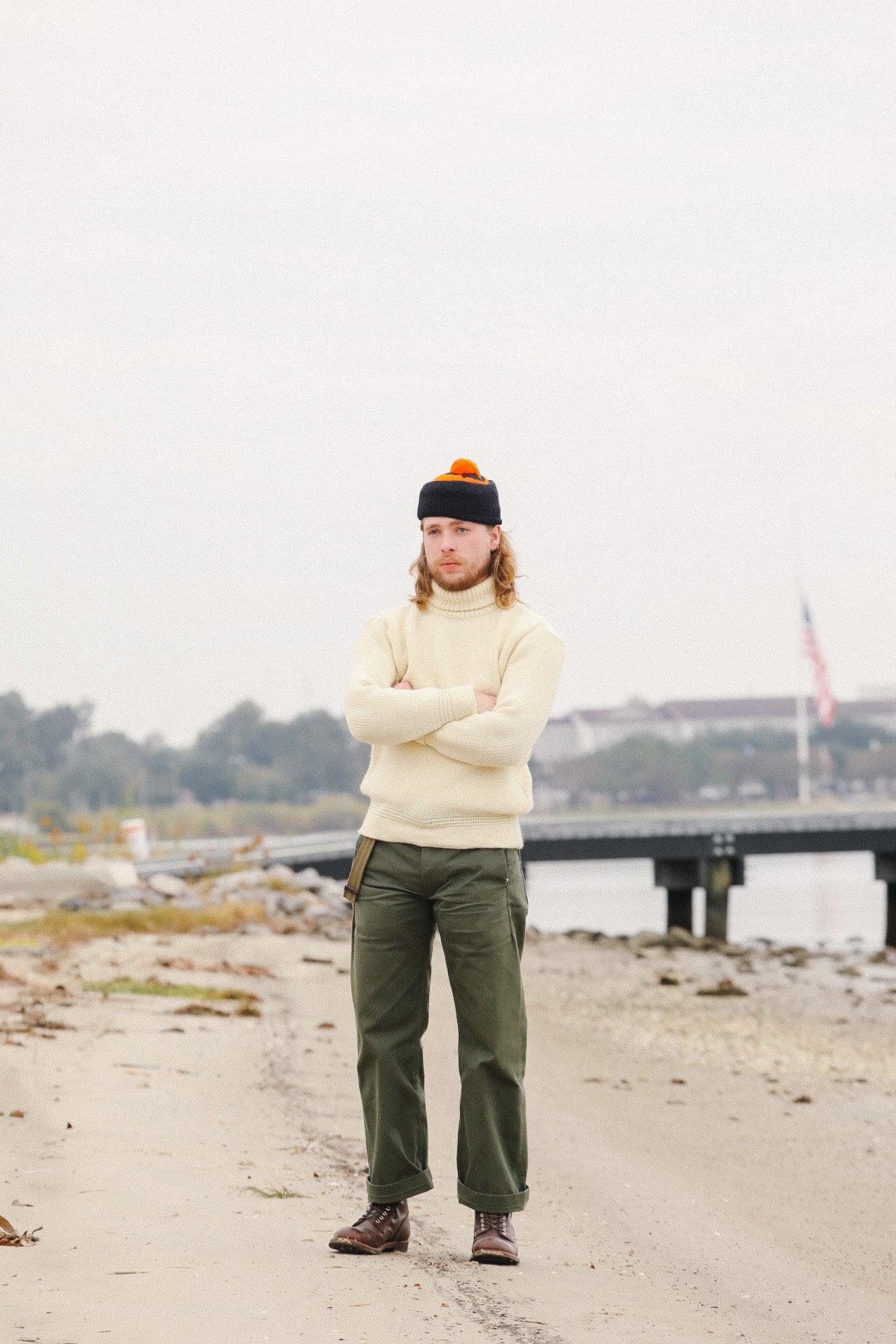 Man wearing cream wool sweater, olive green pants, brown boots, and black beanie standing on sandy beach near pier