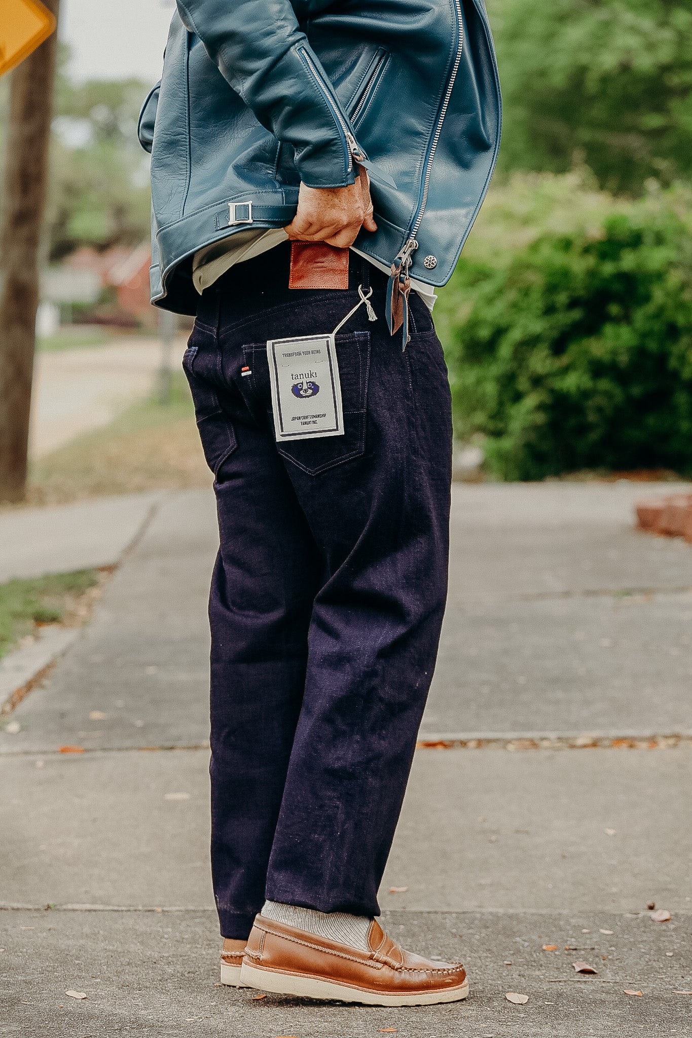 Man wearing dark denim jeans with tanuki label, blue leather jacket, and brown loafers on sidewalk