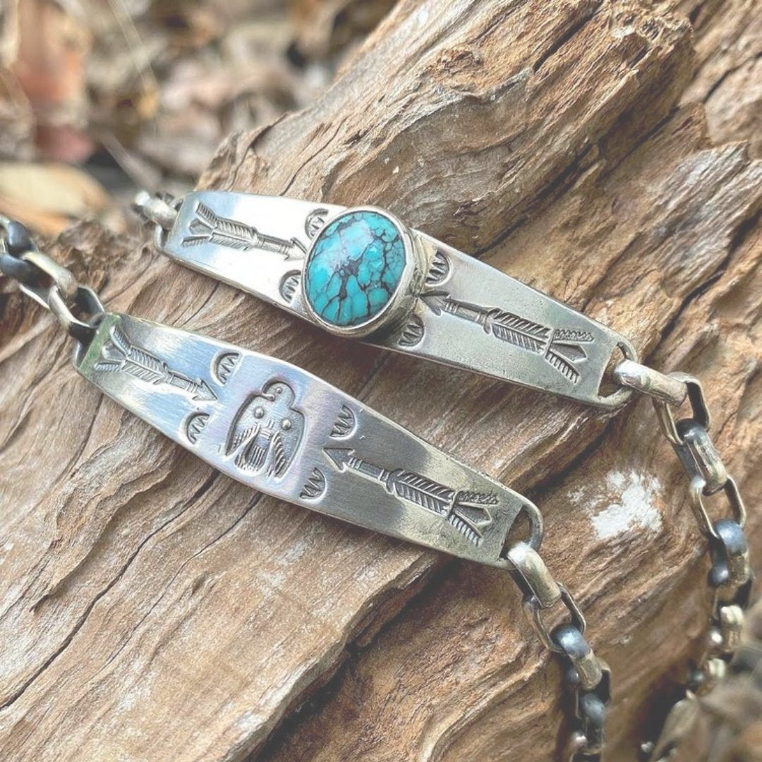 Two handcrafted silver bracelets with engraved arrow and bird designs, one featuring a turquoise stone, on rustic wood