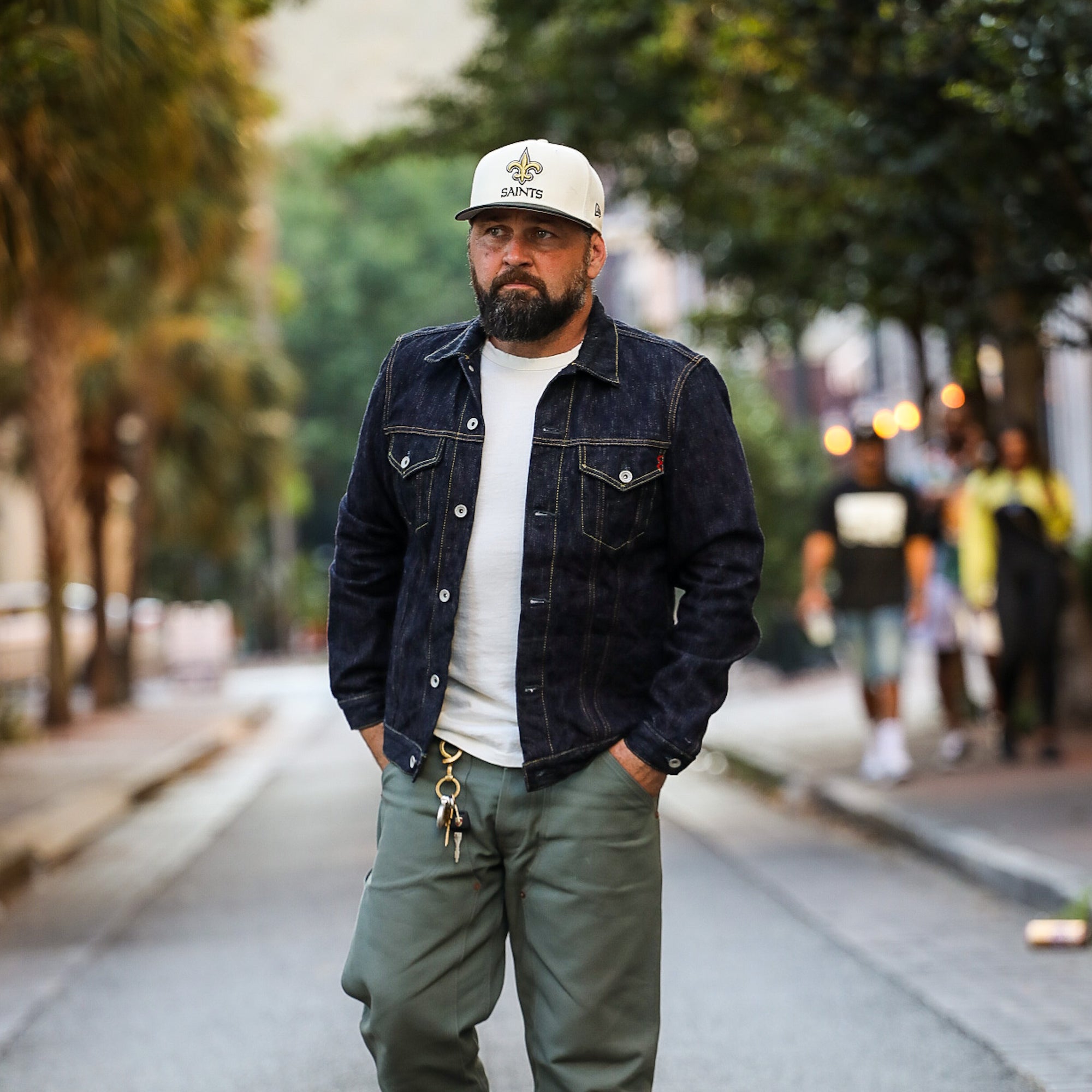Man wearing dark raw denim jacket, white t-shirt, olive pants, and white Saints cap walking on city street