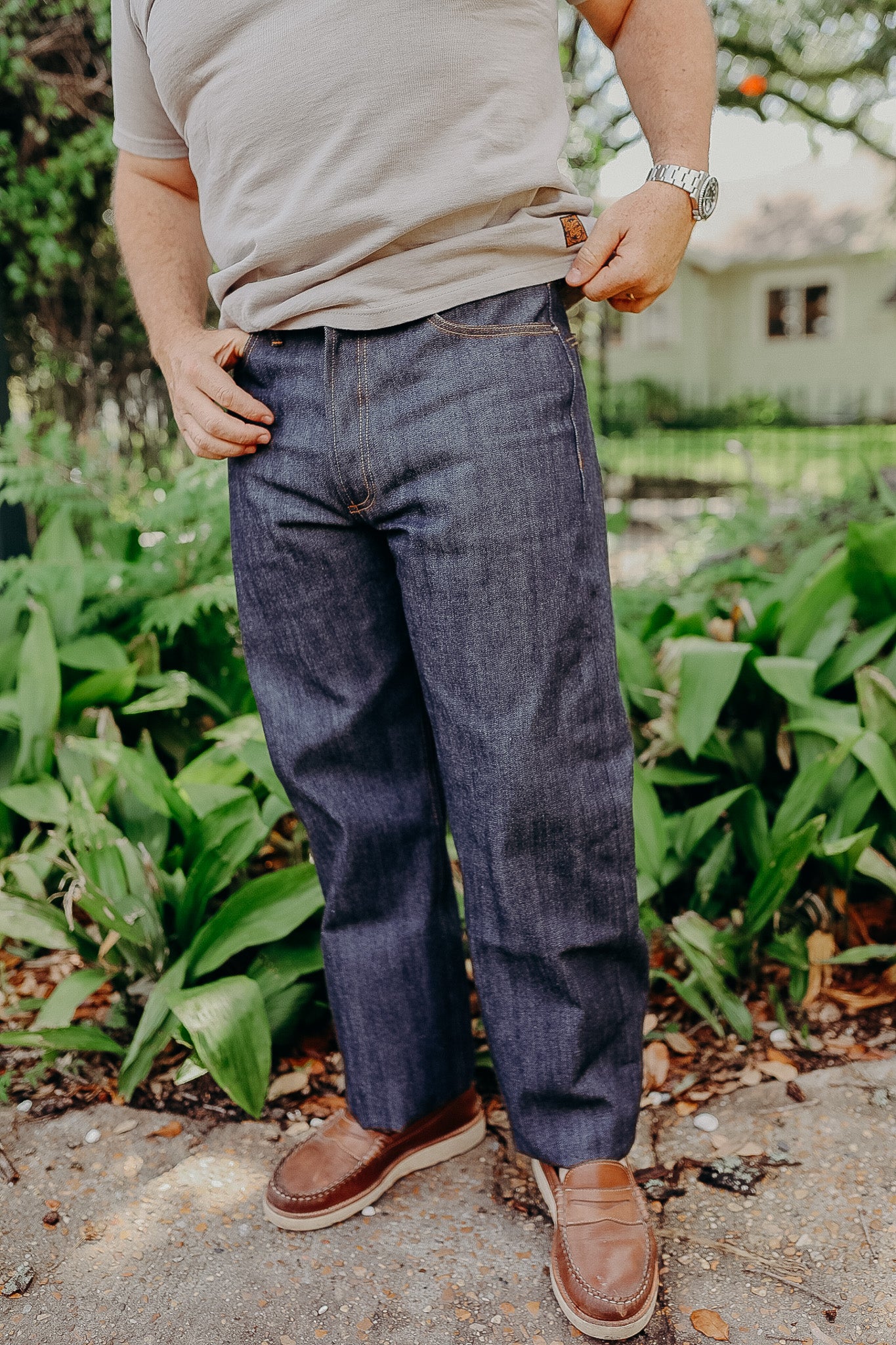 Man wearing straight-cut selvedge raw denim jeans and brown loafers outdoors with greenery background