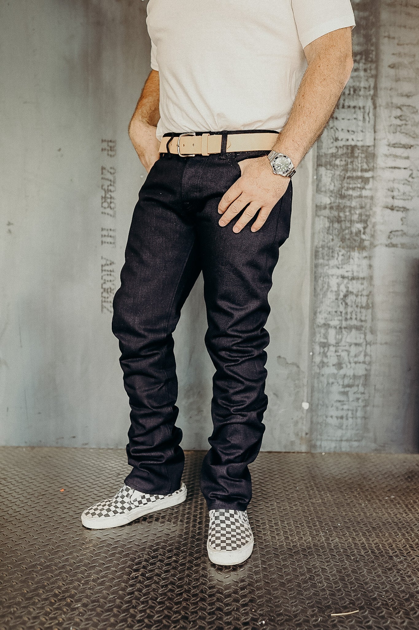 Man wearing slim fit dark selvedge denim jeans, beige belt, checkerboard slip-on shoes, and white t-shirt