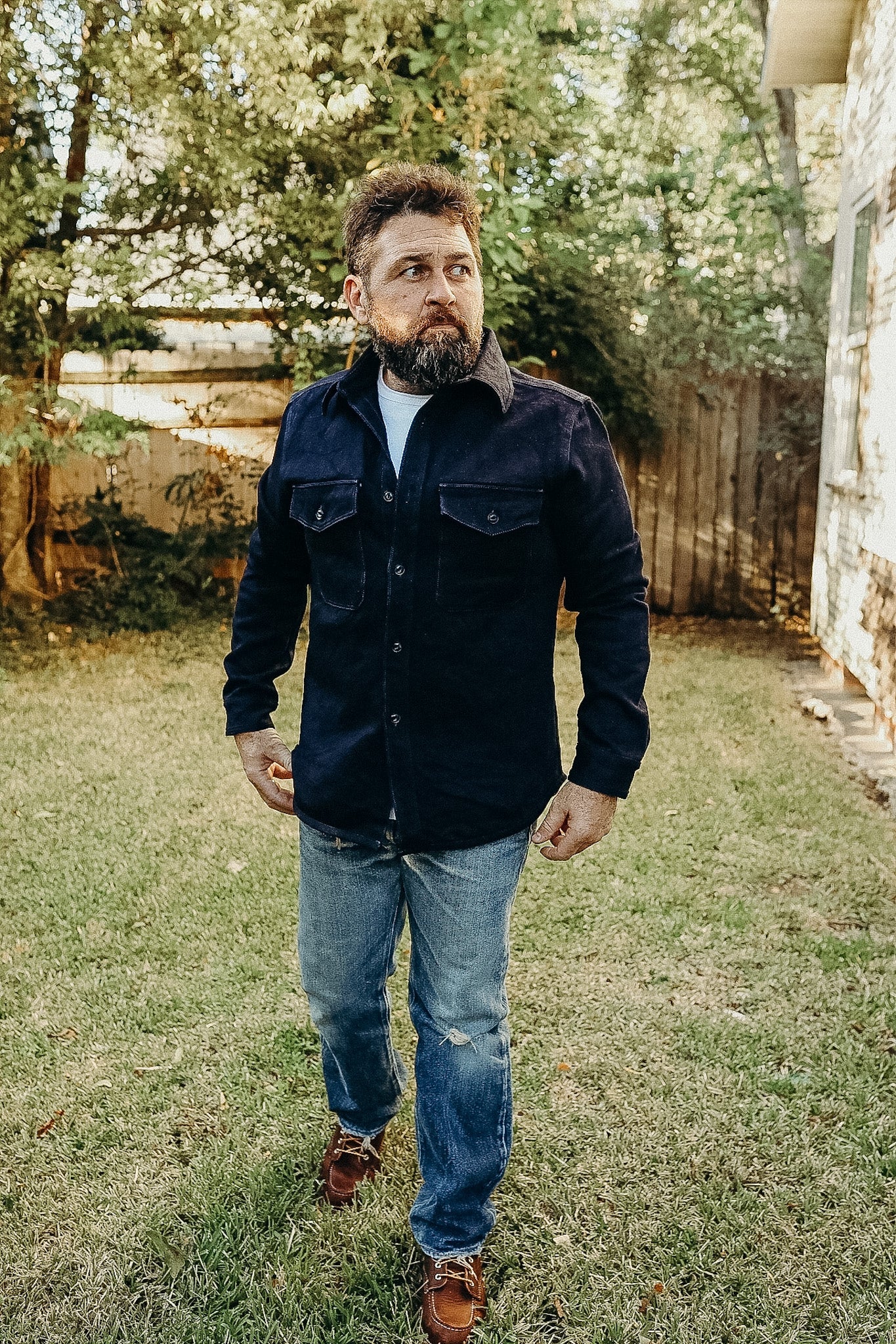 Man wearing indigo rope dye tricotine shirt with chest pockets, blue jeans, and brown leather shoes in a backyard
