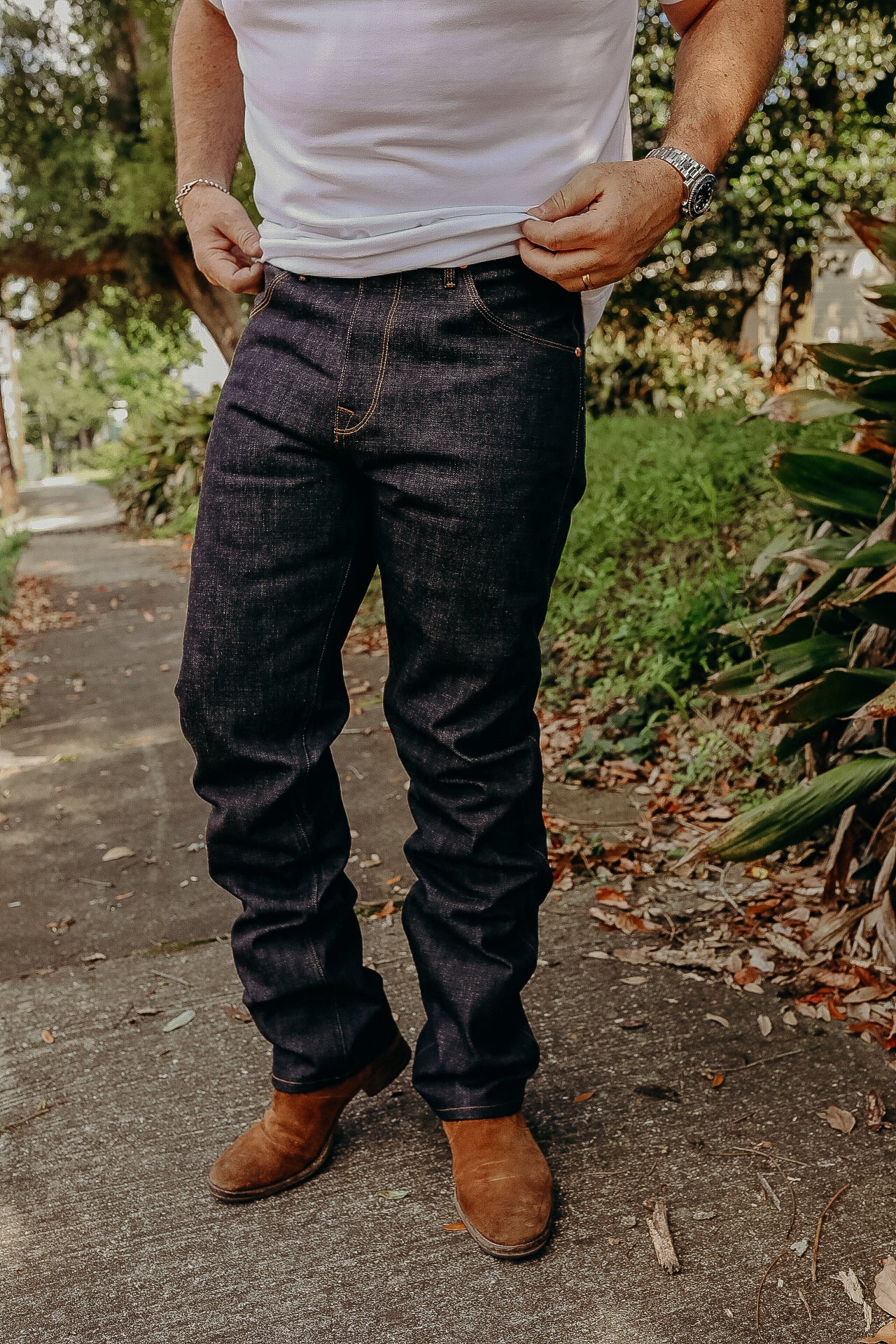 Man wearing dark raw denim western cut jeans and brown leather boots on sidewalk with greenery background