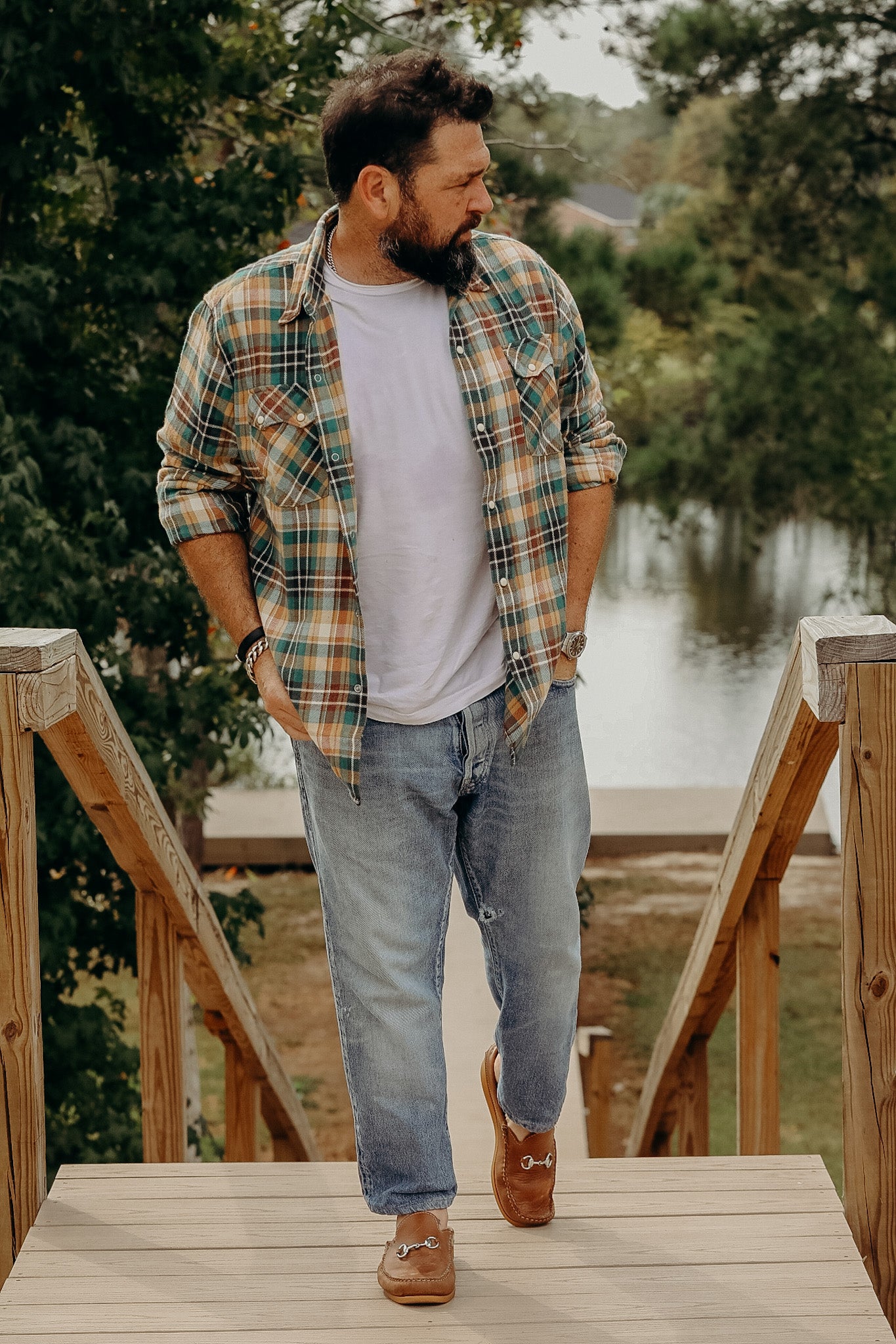 man wearing Wythe Americana Western plaid shirt over white tee and jeans on wooden stairs near lake