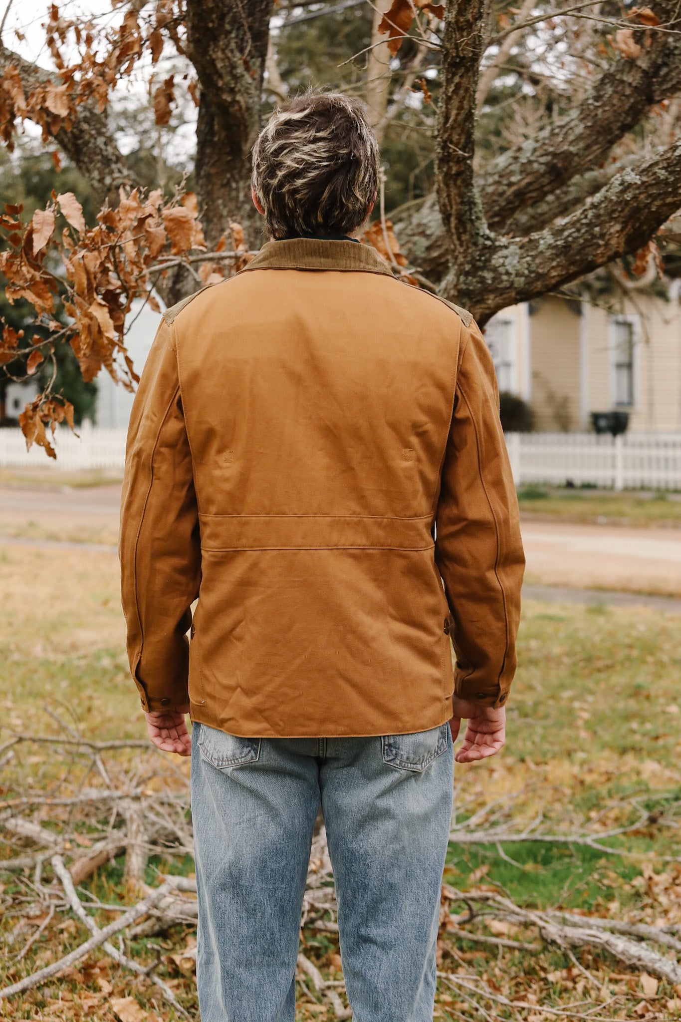 Person wearing a brown jacket standing outdoors with trees and grass in the background