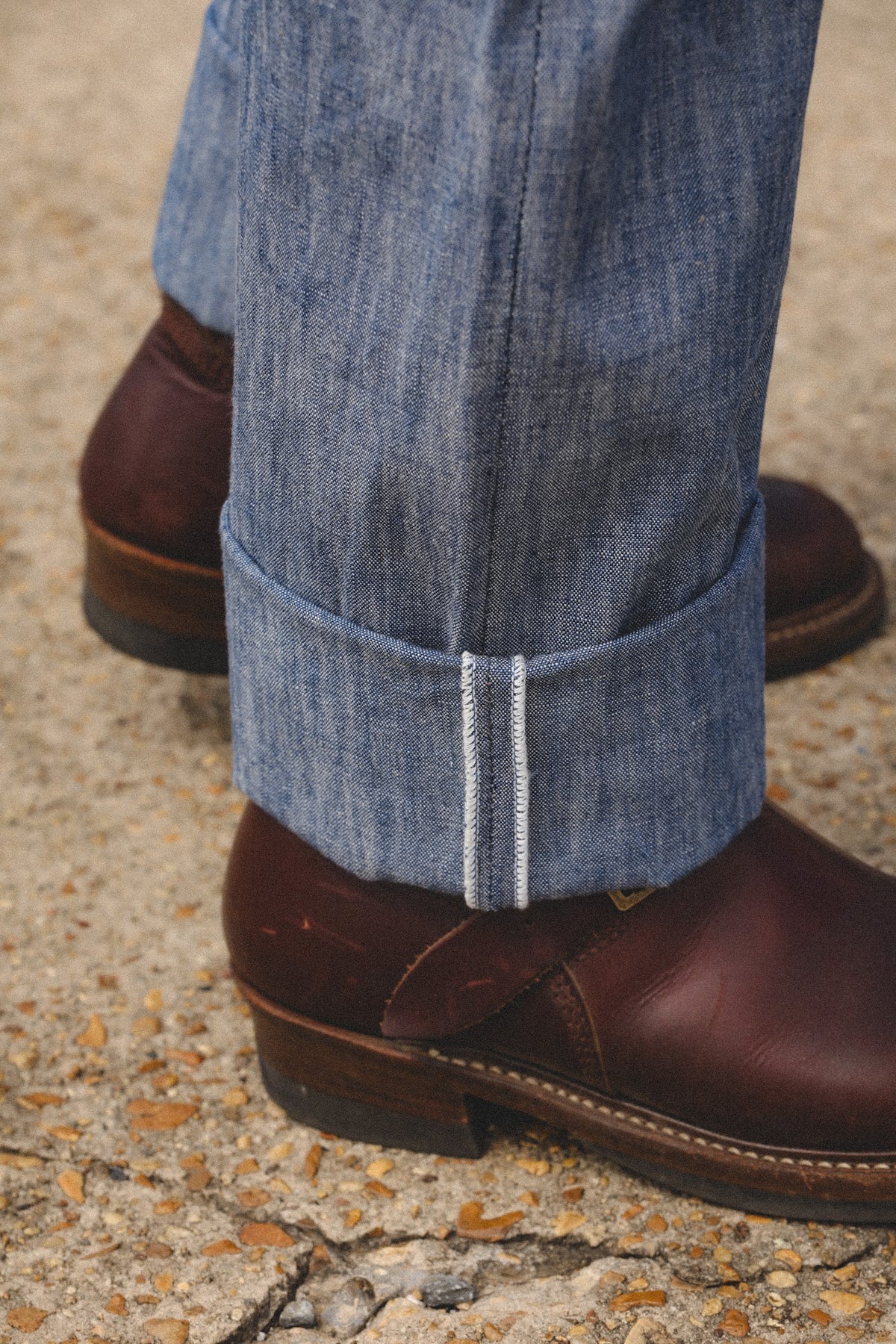 Close-up of a person's legs wearing blue denim jeans with a rolled cuff and dark brown leather boots on a textured concrete ground.