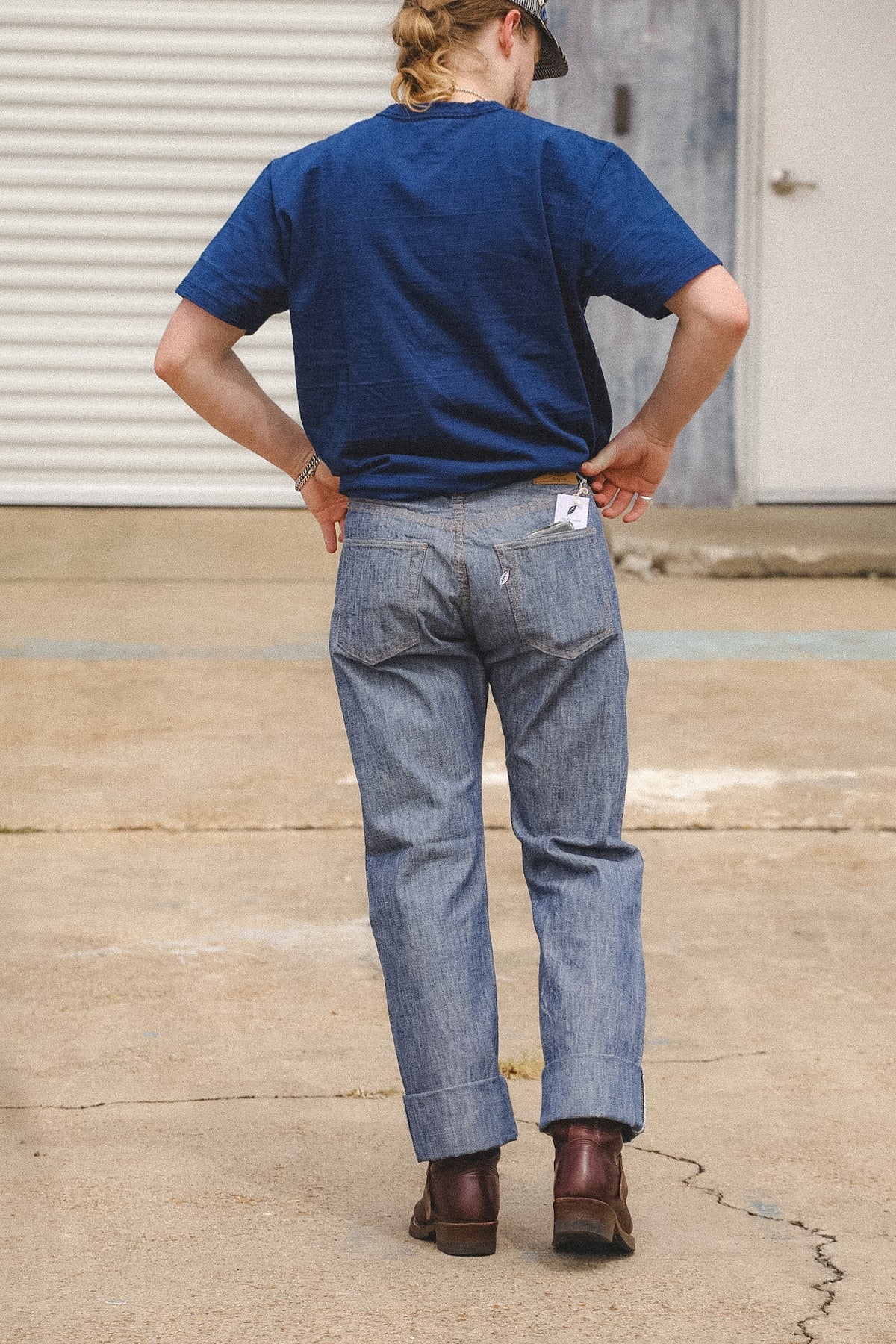 A person viewed from behind wearing a navy blue t-shirt, light-wash blue jeans, and brown boots, standing on concrete in front of a garage door.
