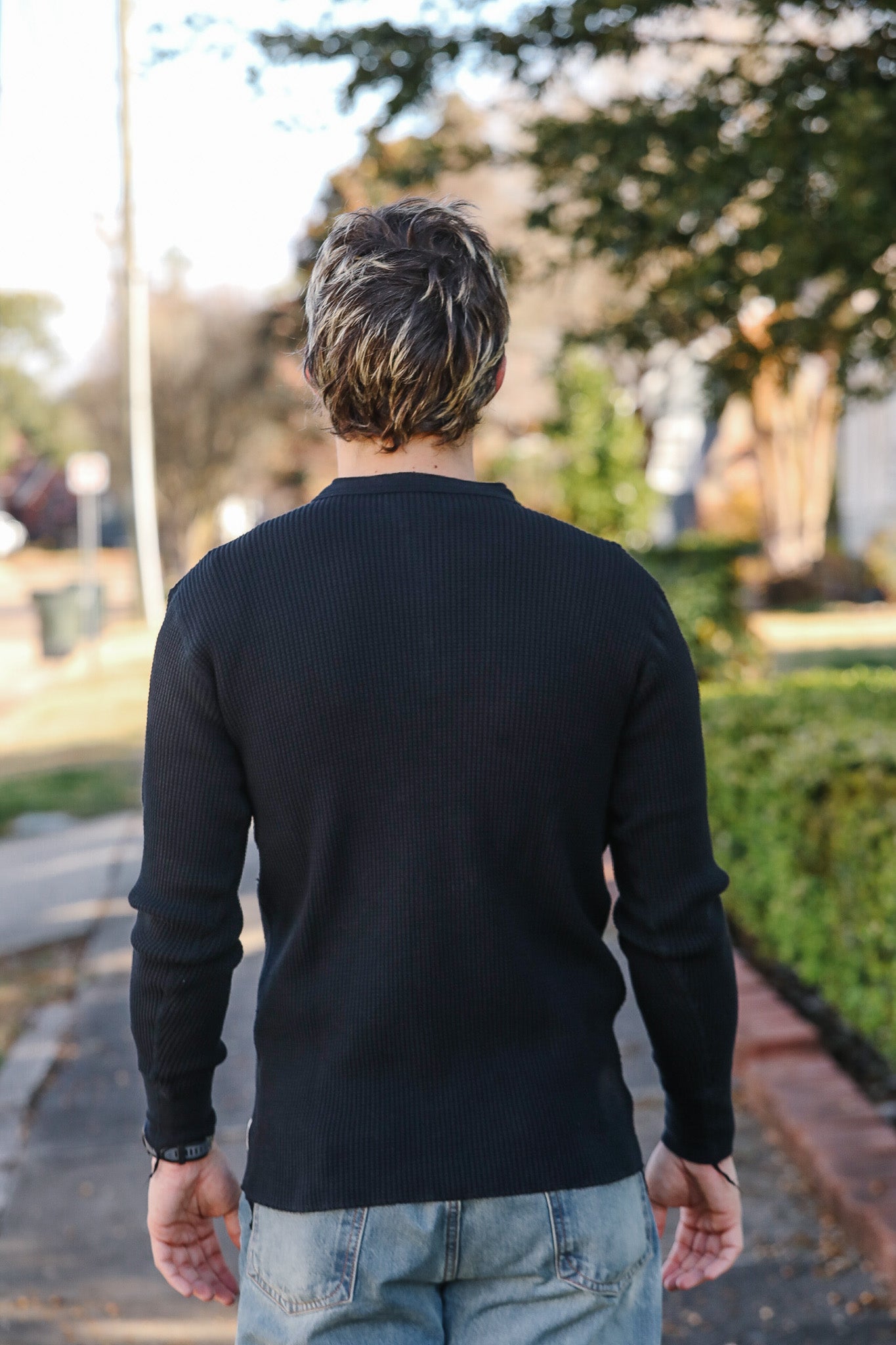 Person wearing a black sweater and light blue jeans walking away on a path with trees and grass in the background.
