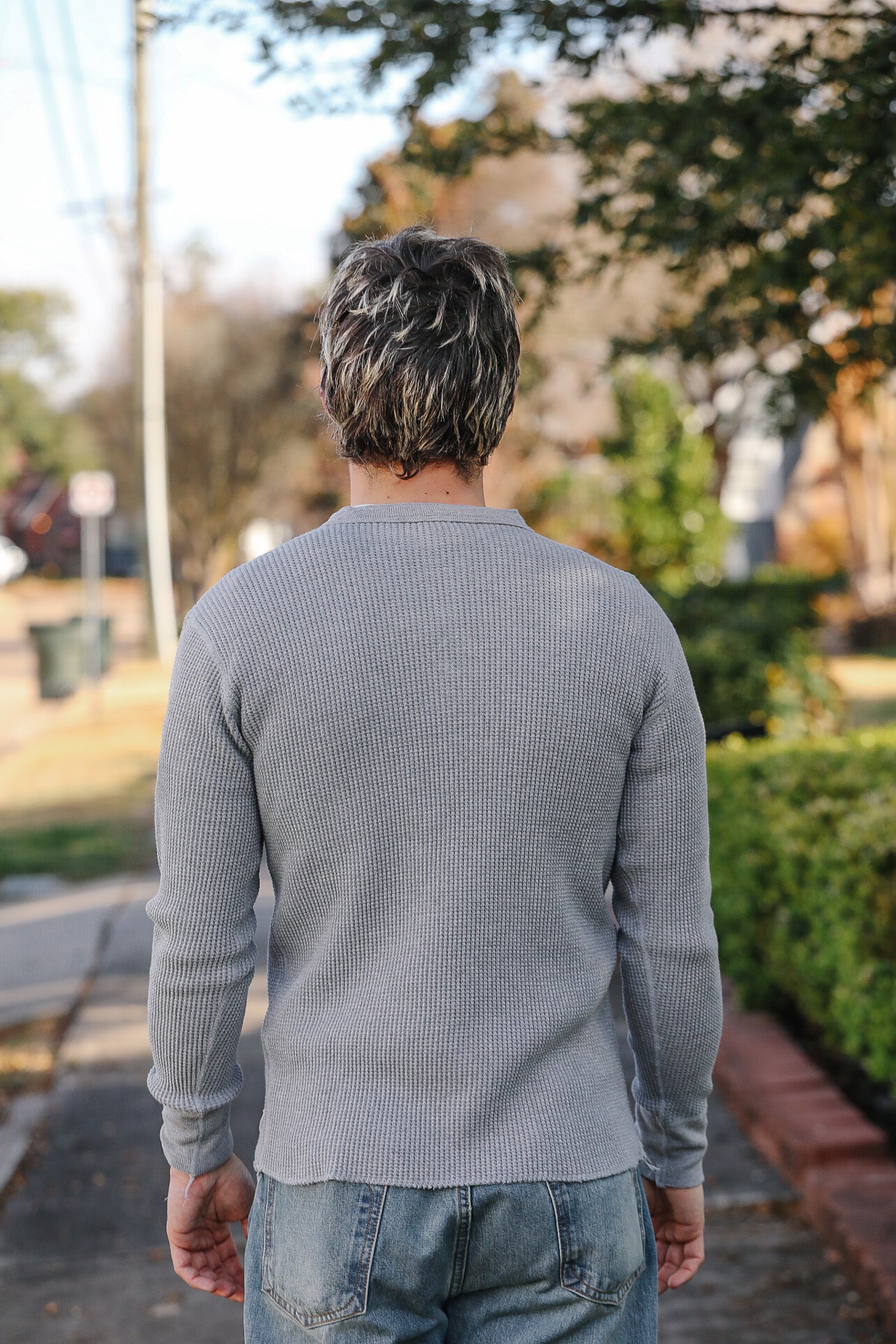 Man wearing a gray sweater and jeans standing on a street with trees and a house in the background.
