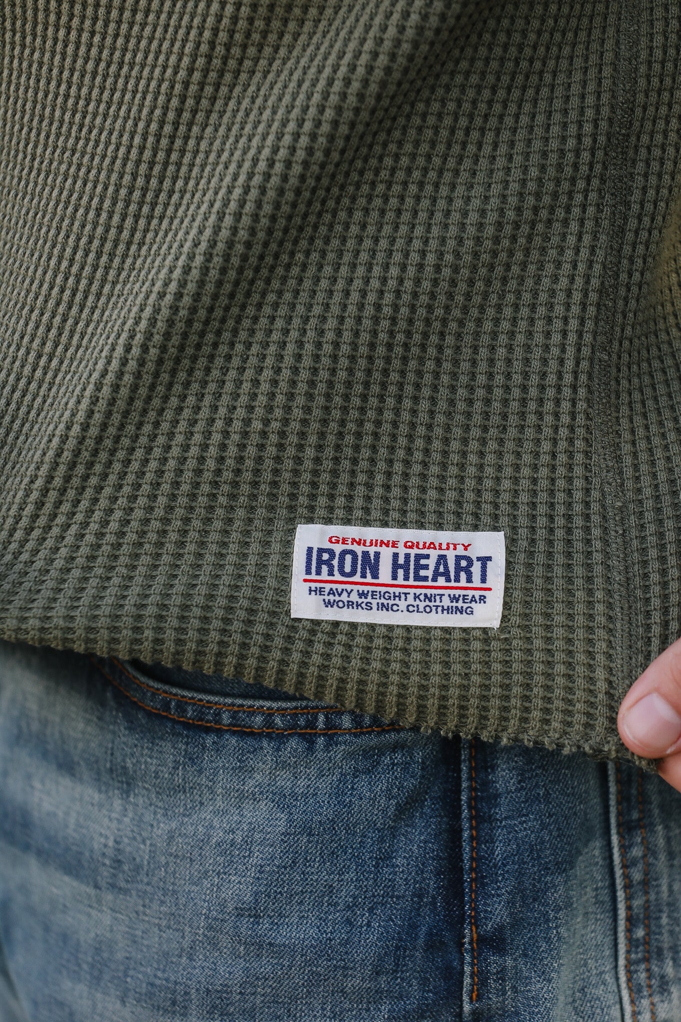 Close-up of a green textured shirt with an Iron Heart label on a person wearing blue jeans.