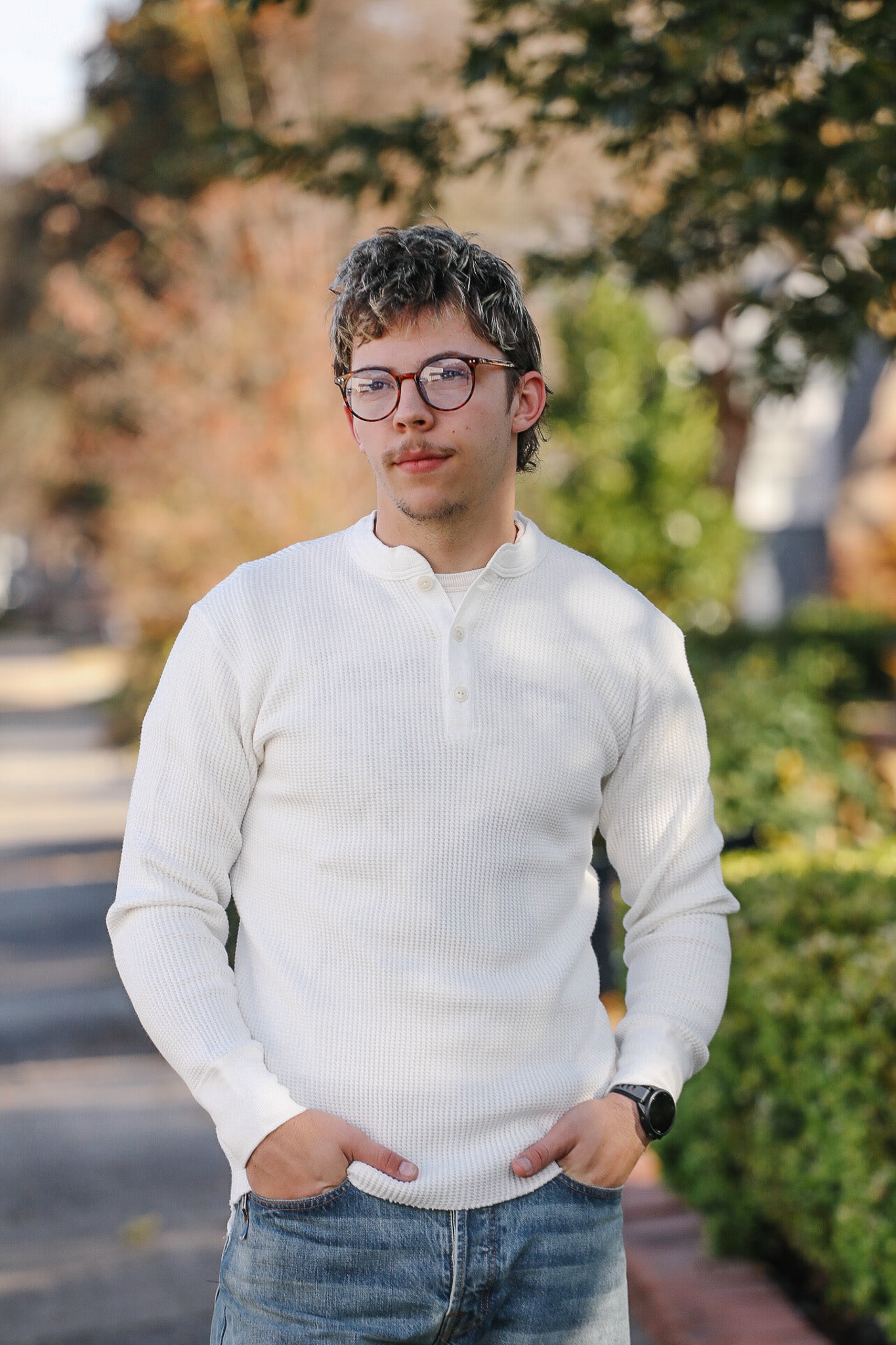 Person wearing a white sweater and glasses outdoors with greenery in the background