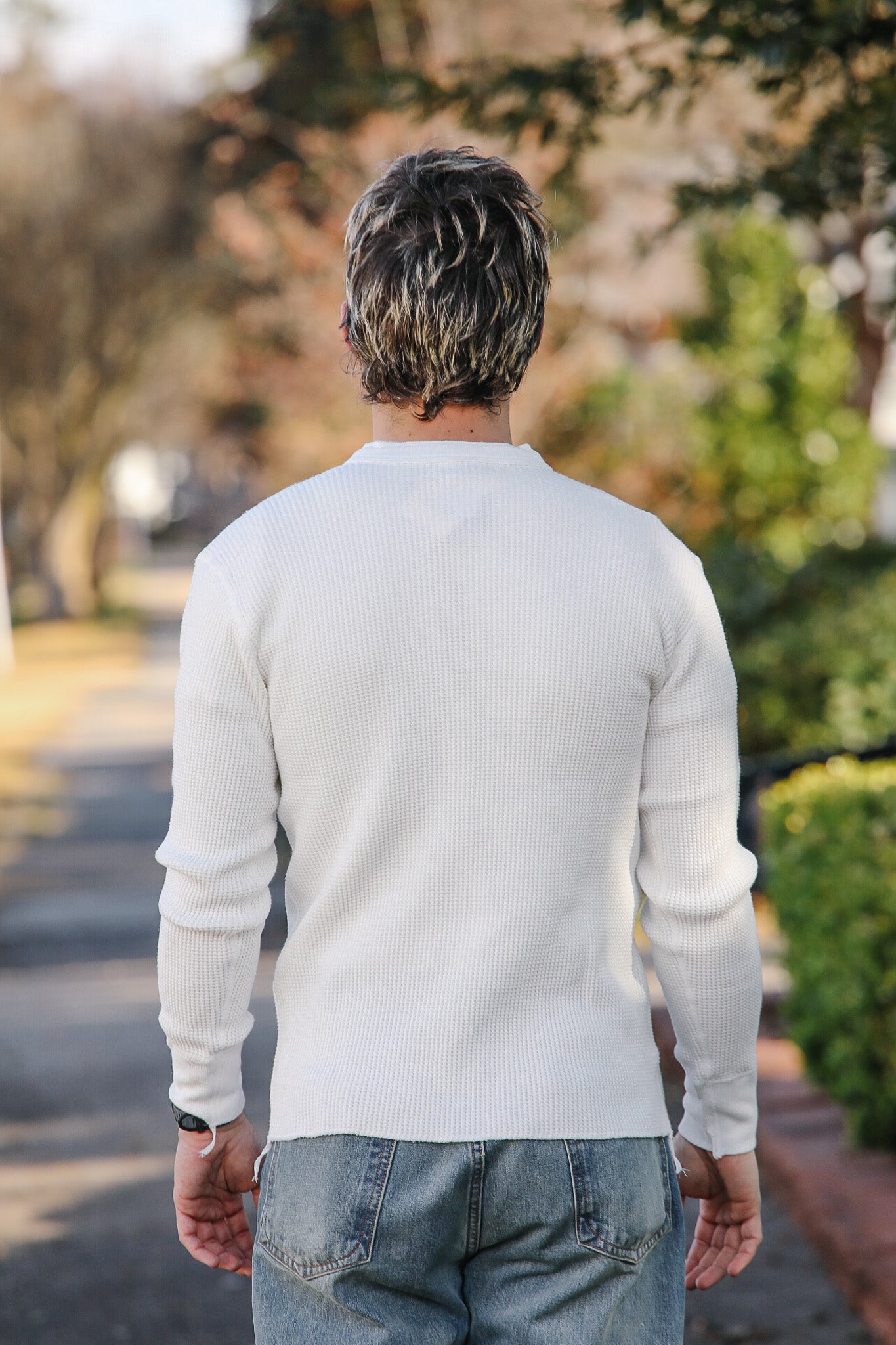 Man wearing a white sweater and blue jeans walking away from the camera with trees and road in the background.