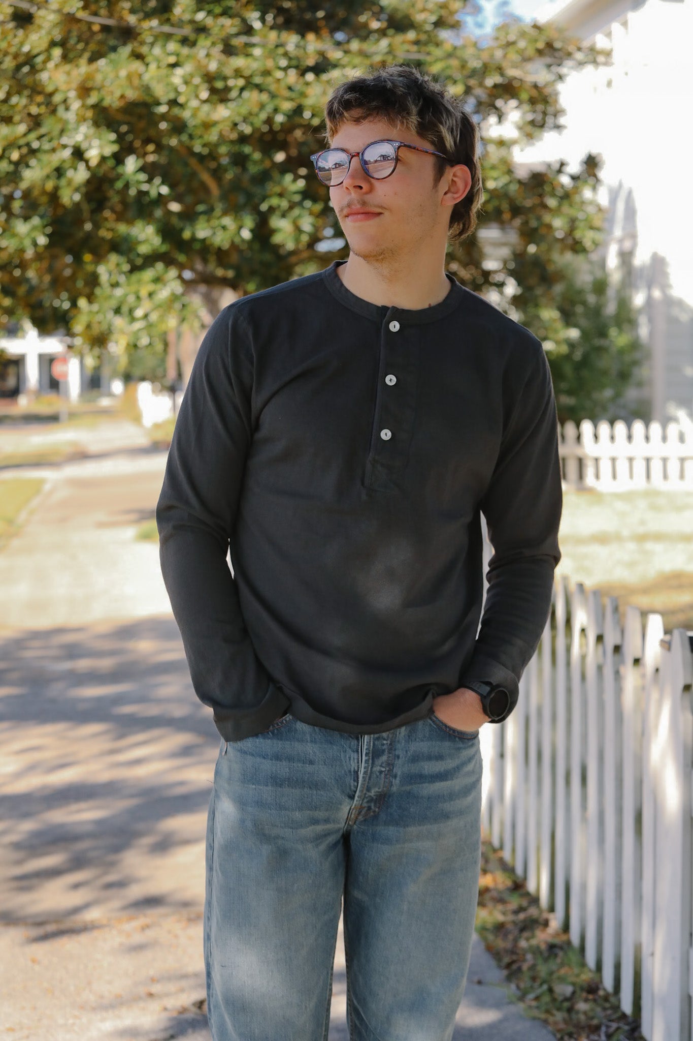 Person wearing a black long-sleeve shirt and blue jeans standing outdoors with trees and a white fence in the background.