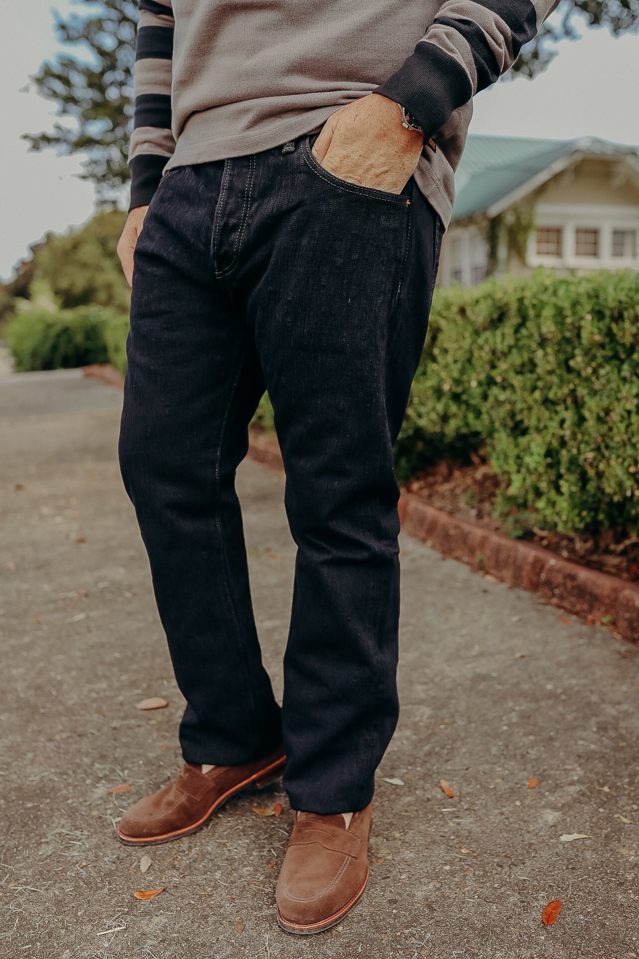 Man wearing dark denim jeans and brown suede loafers standing on outdoor path with greenery and house in background