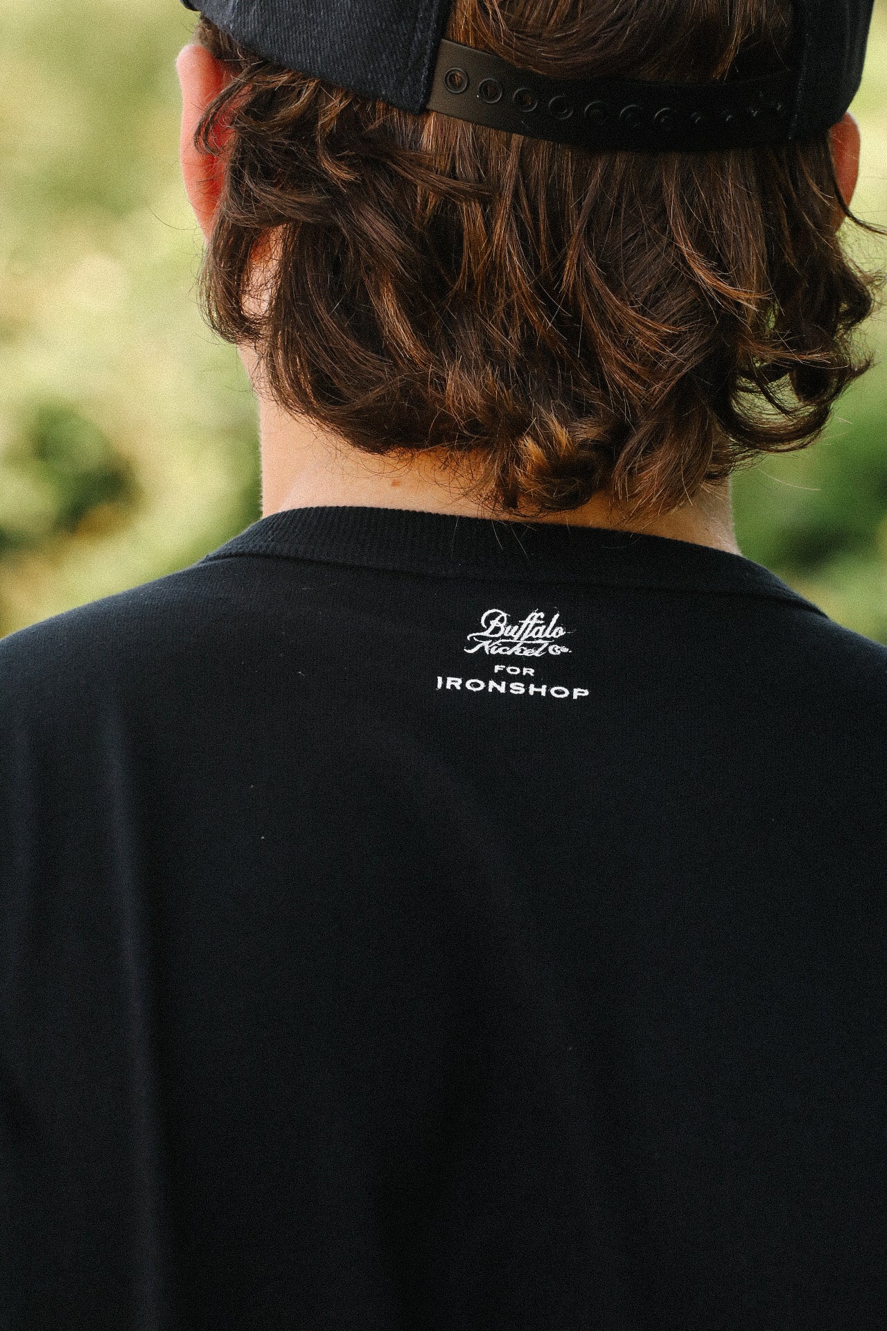 Back view of person wearing black Buffalo Nickel Coffin Loopwheel T-shirt with white logo above collar outdoors