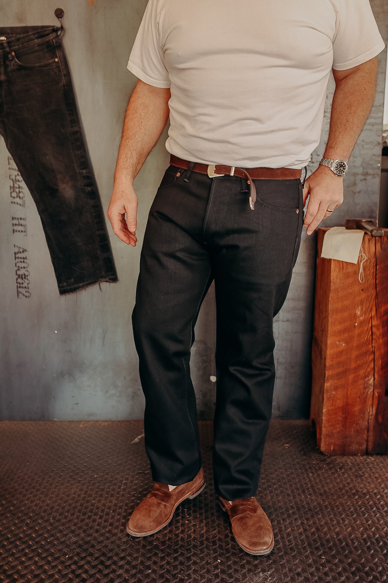 Man wearing dark denim jeans with brown belt and brown suede shoes in industrial setting