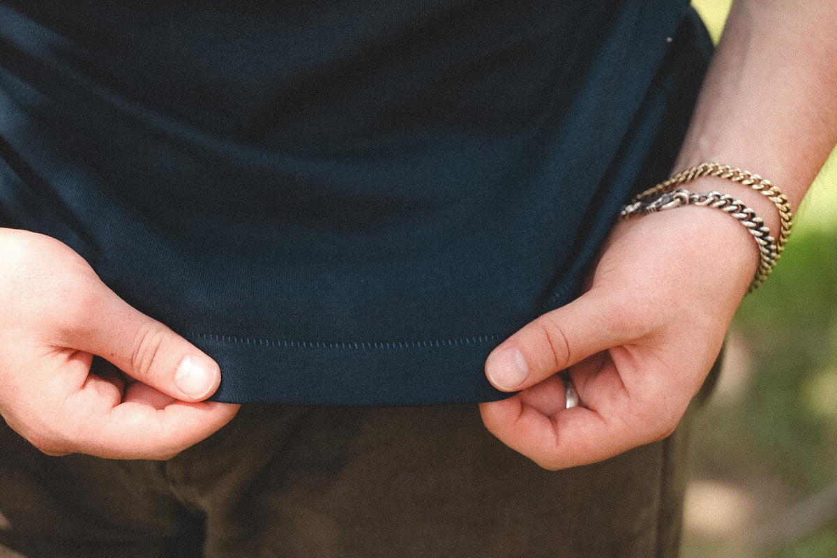Close-up of a person's hands pulling up the hem of a dark navy blue t-shirt, with gold chain bracelets visible on the wrist against a blurred outdoor background.