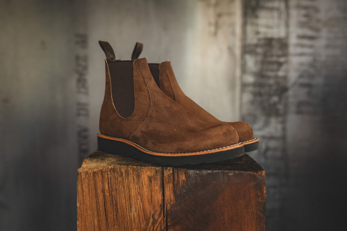 A pair of brown suede Chelsea boots with dark elastic side panels and pull tabs, resting on a rustic wooden block against a blurred gray background.