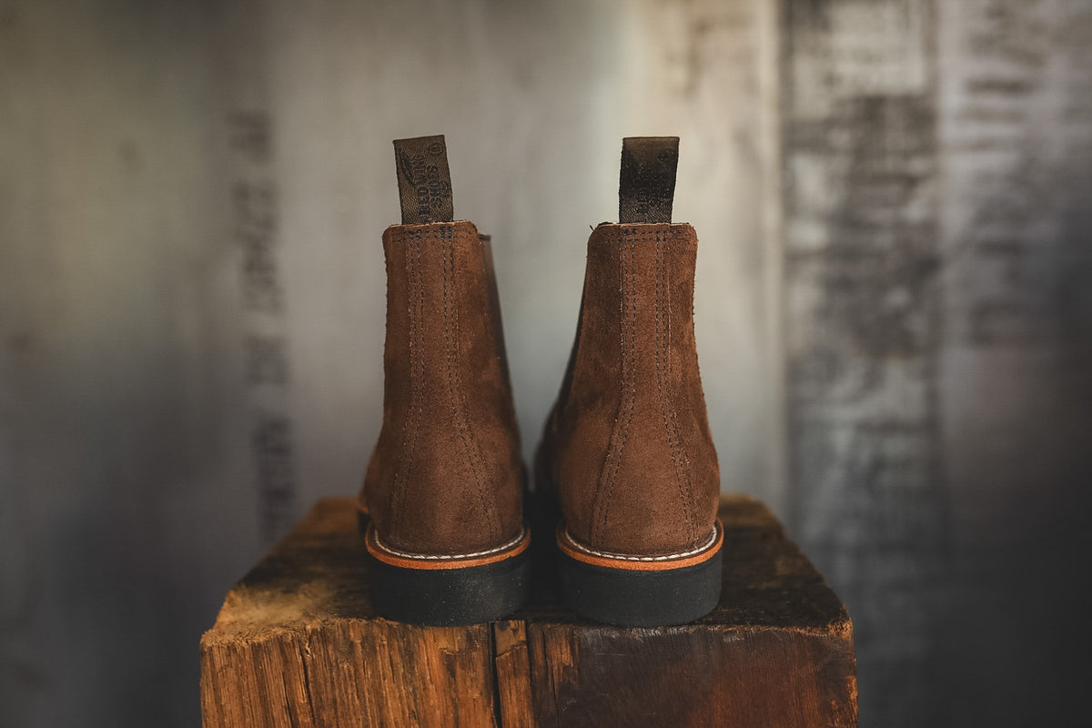 A pair of brown suede Chelsea boots shown from the back, resting on a rustic wooden block against a blurred gray background.