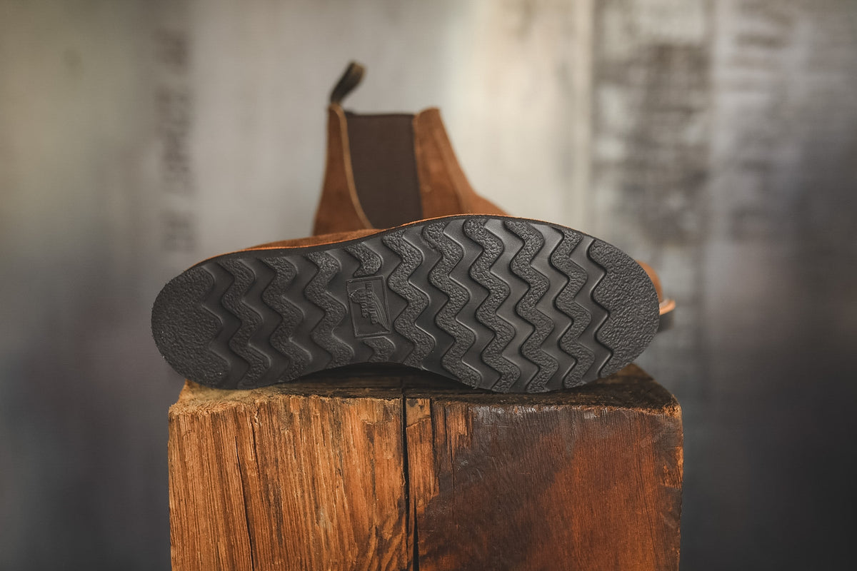 A close-up shot of a brown leather ankle boot shown from the bottom, displaying its textured black zigzag treaded outsole resting on a rustic wooden block against a blurred gray background.
