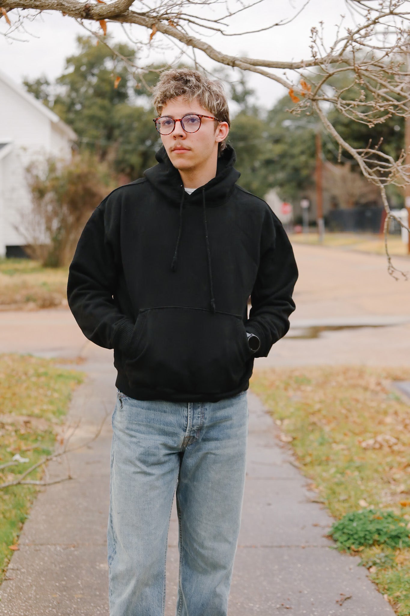 Person wearing a black hoodie and jeans standing on a sidewalk with trees and houses in the background.