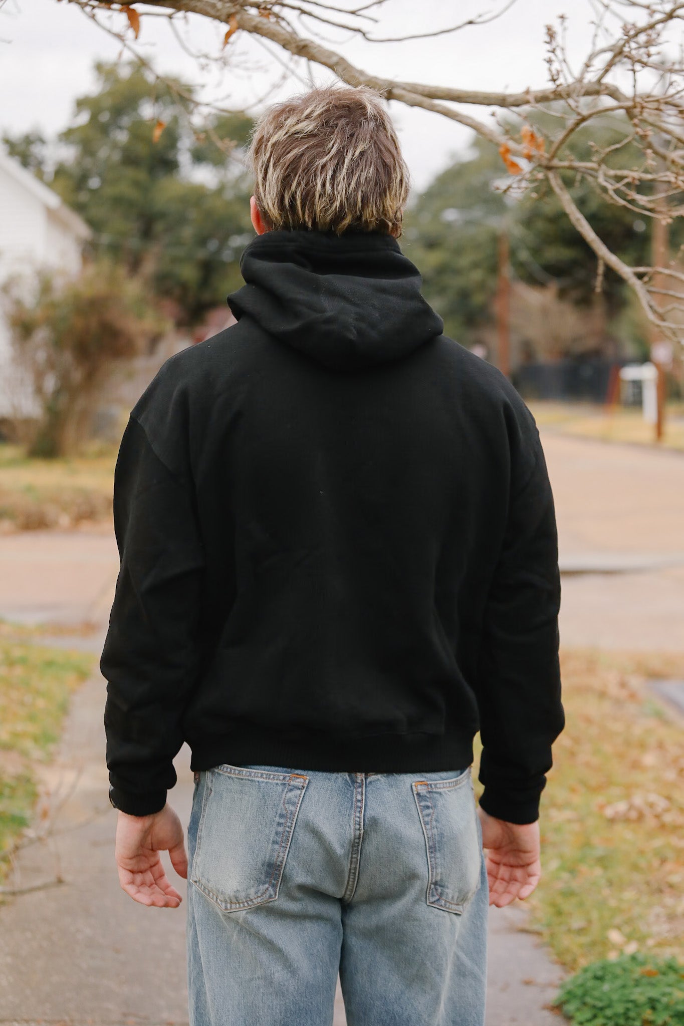 Person wearing a black hoodie and light blue jeans walking away from the camera in a suburban setting.