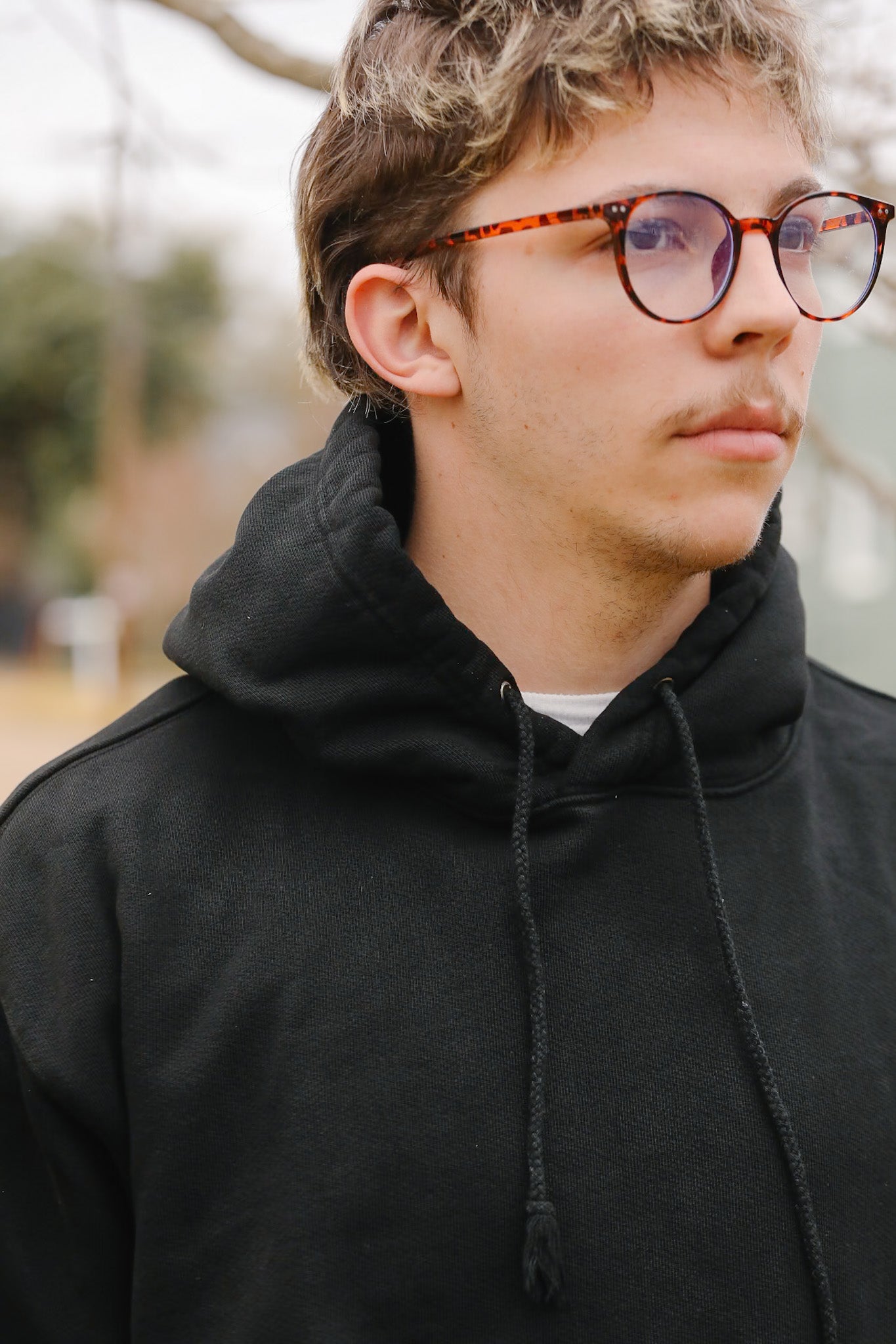 Person wearing a black hoodie and glasses with a blurred outdoor background