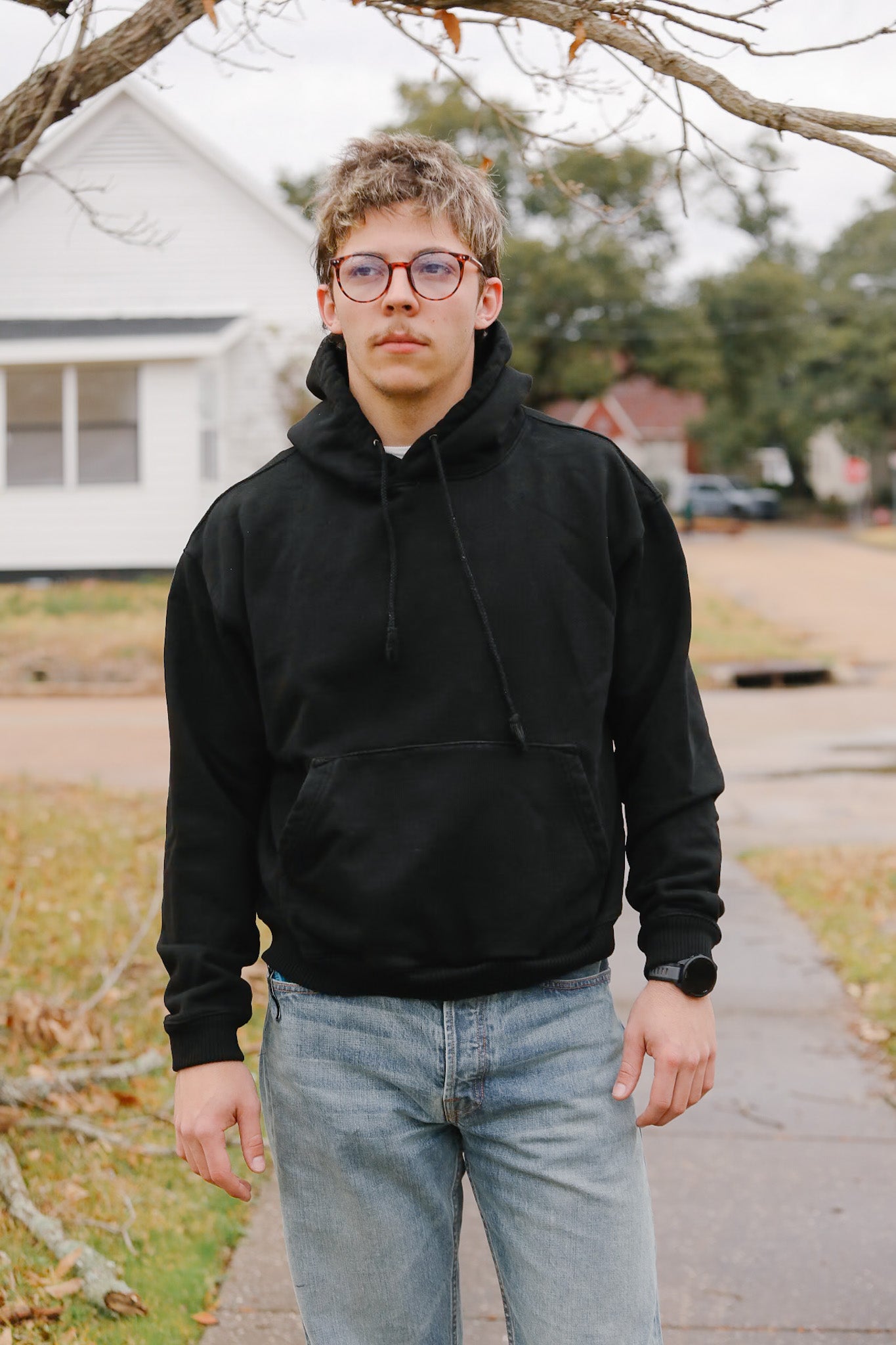 Person wearing a black hoodie and glasses standing on a sidewalk with a house in the background