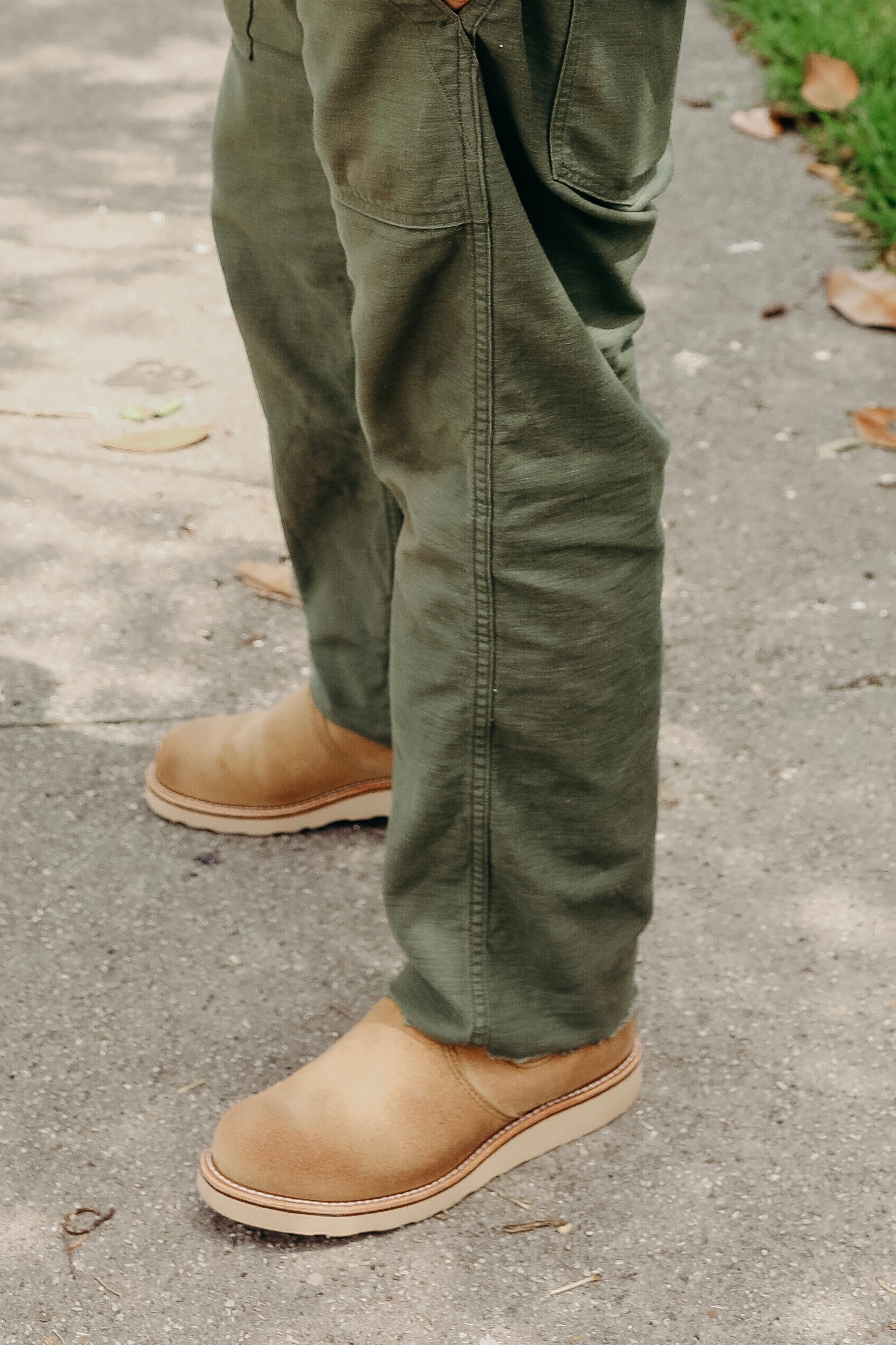 Close-up of a person wearing olive green work pants and tan slip-on boots standing on a concrete path