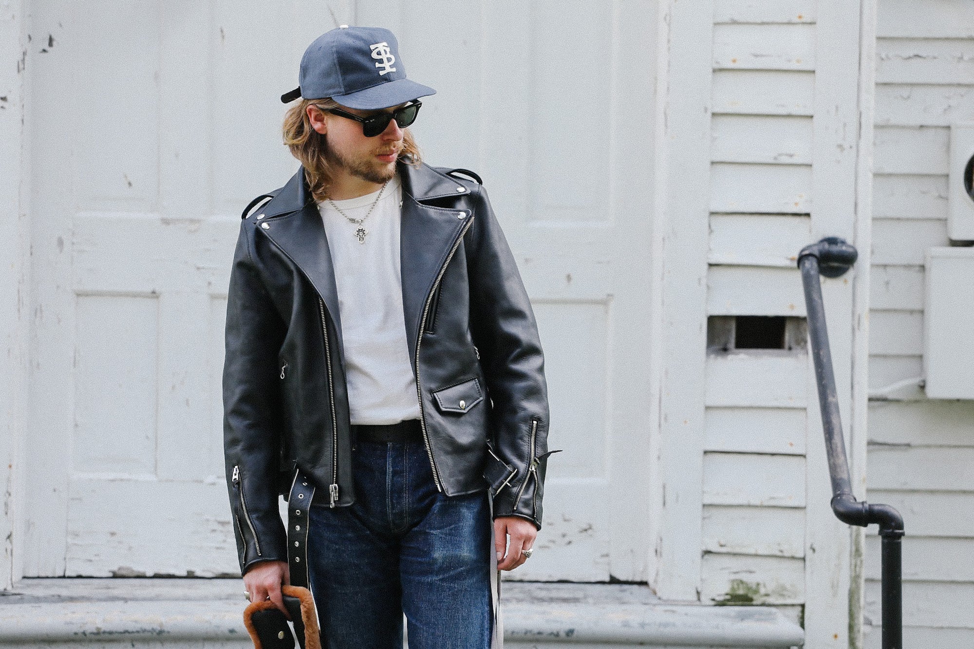 Man wearing black Sumi Dyed Double Riders leather jacket, white t-shirt, blue jeans, and blue cap, standing outdoors