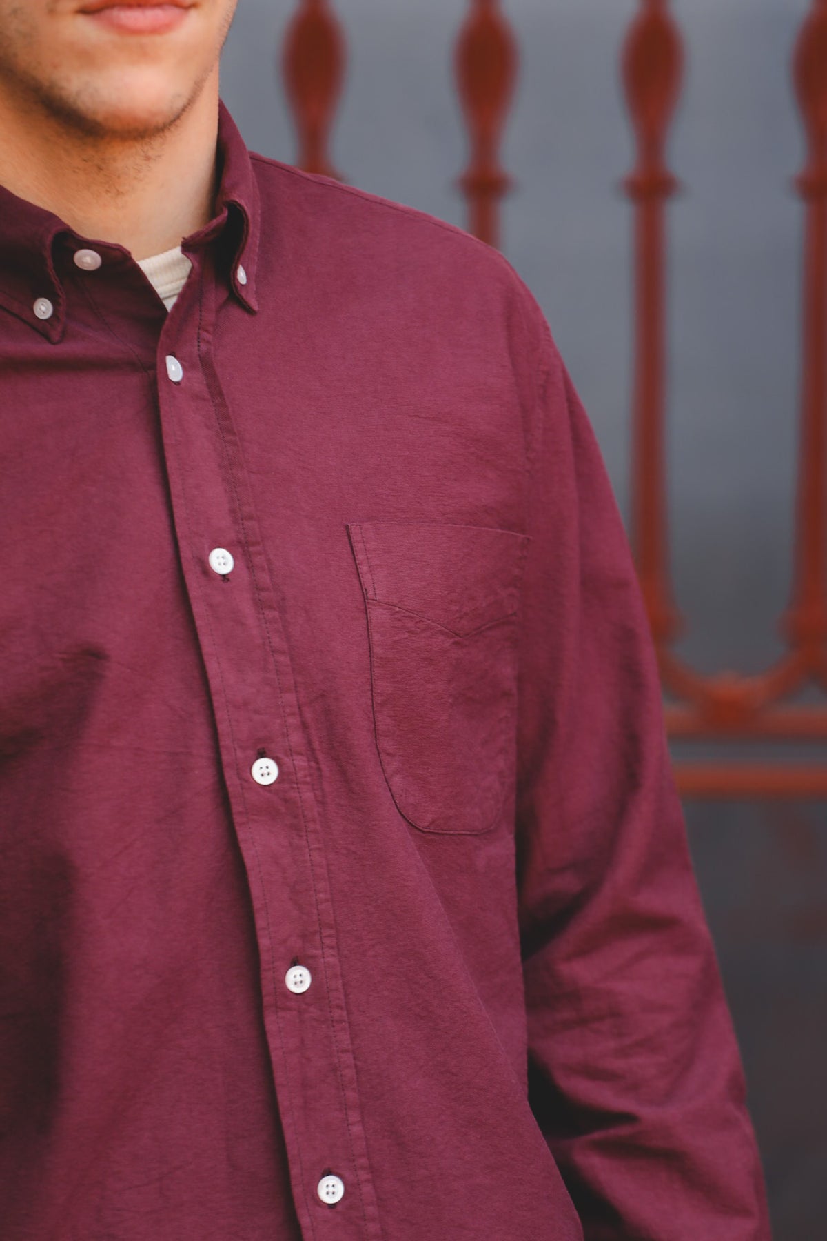 A close-up of a man wearing a burgundy long-sleeve button-down shirt with a chest pocket, shown from the neck down against a blurred outdoor background.