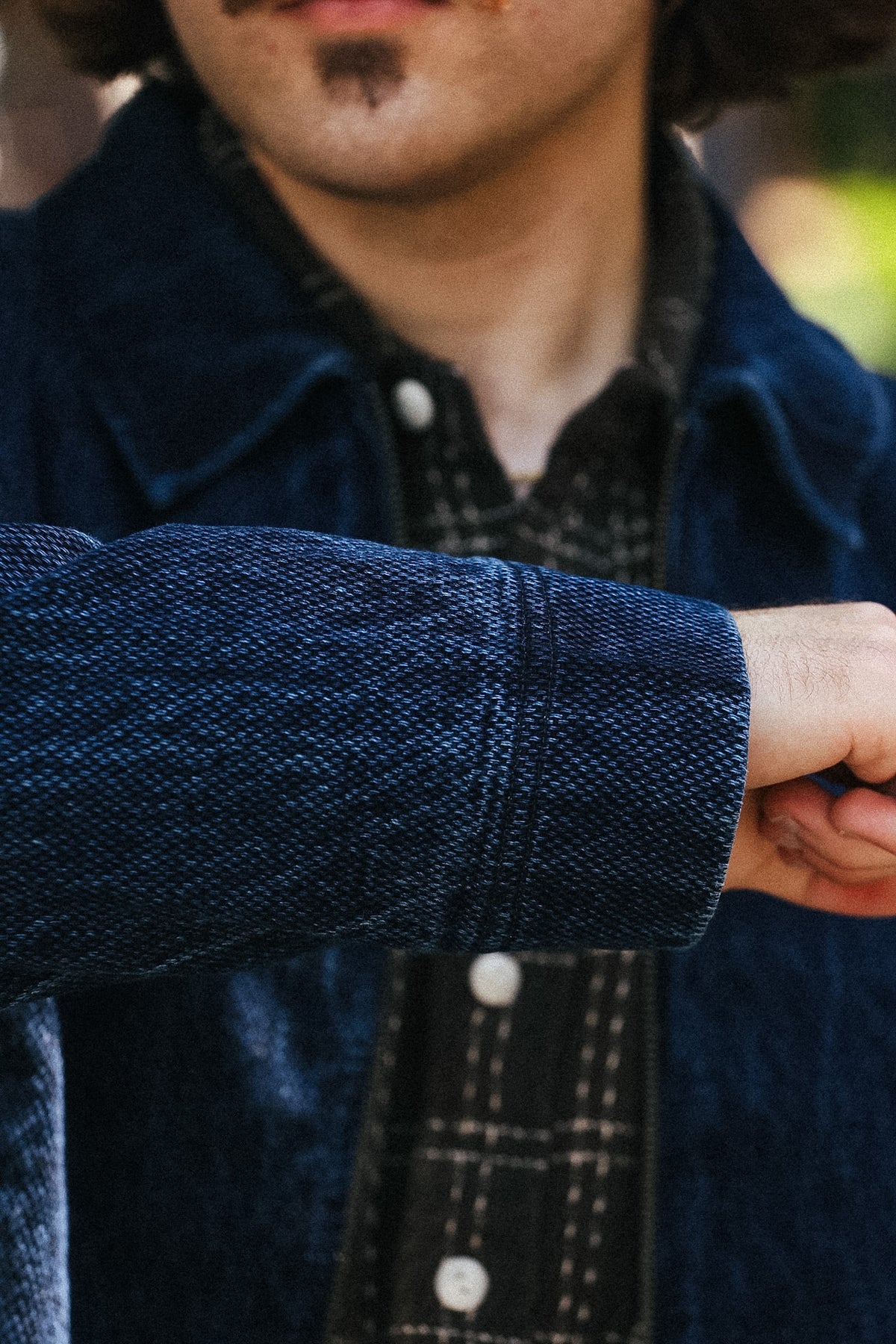 Close-up of a person wearing a dark blue textured corduroy jacket over a black and white plaid button-up shirt, with the person's chin, mouth, and hand visible.