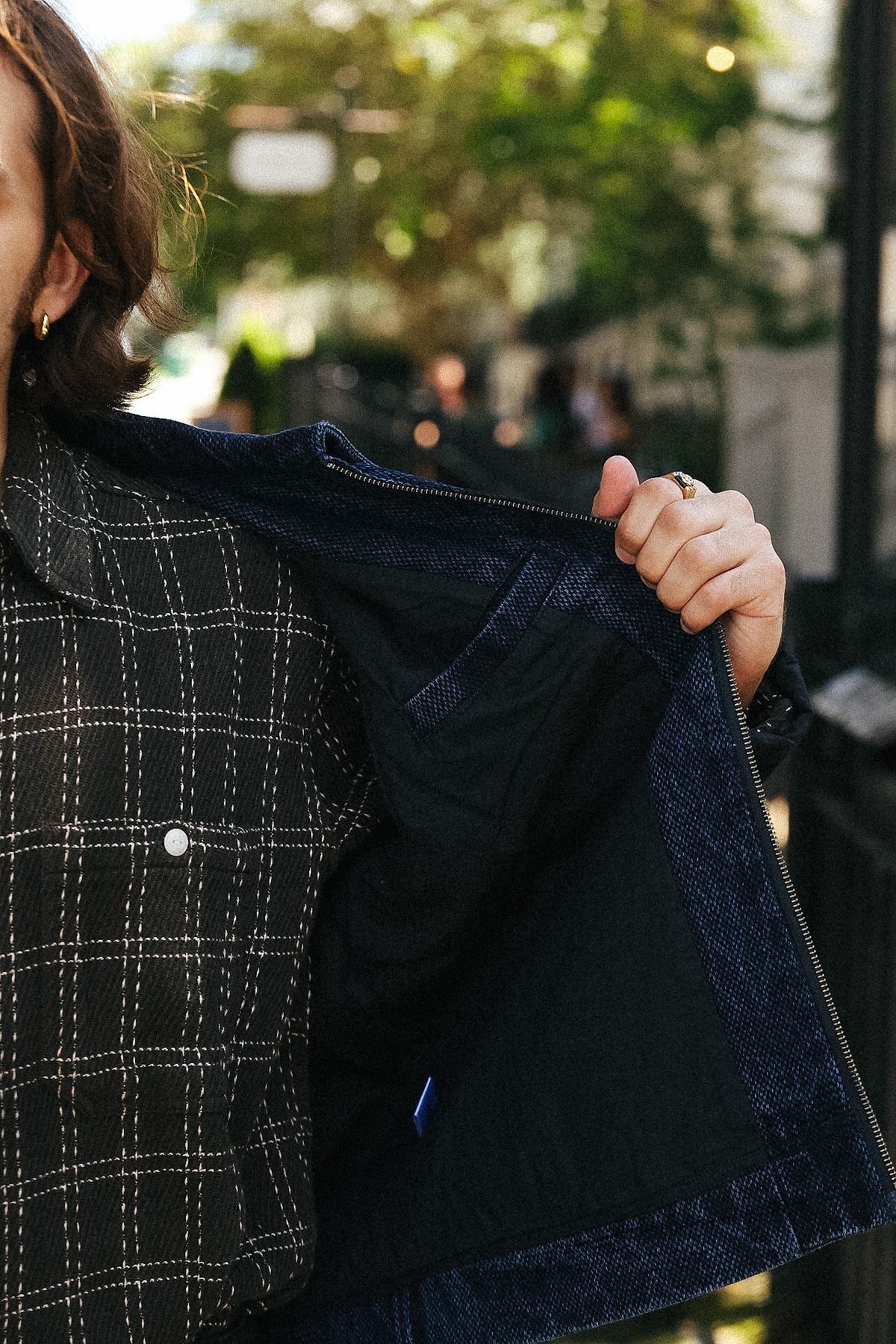 A close-up lifestyle shot of a person holding open a dark blue denim jacket to show its interior lining, wearing a black and white plaid button-up shirt. The background is a softly blurred outdoor street scene with green foliage.