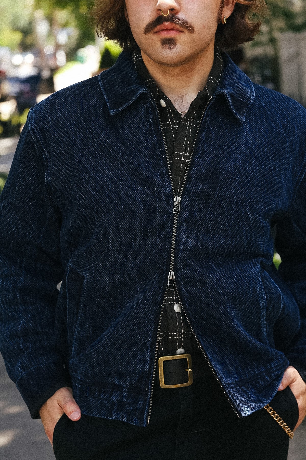 A man wearing a dark navy blue corduroy jacket over a black plaid shirt and black pants with a gold buckle belt, standing outdoors.