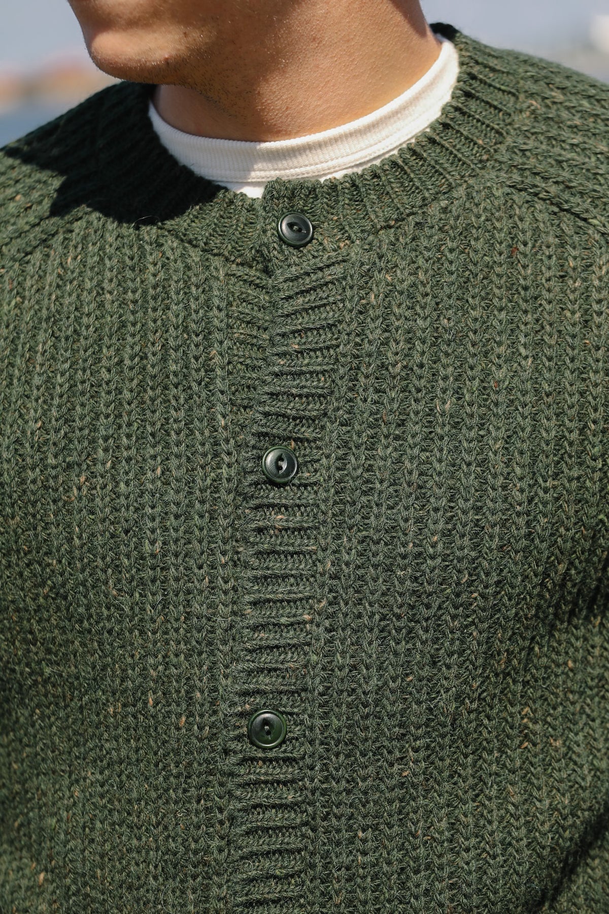 Close-up of a person wearing a textured dark green knit cardigan over a white crewneck shirt, with the cardigan featuring a ribbed button placket and dark buttons.