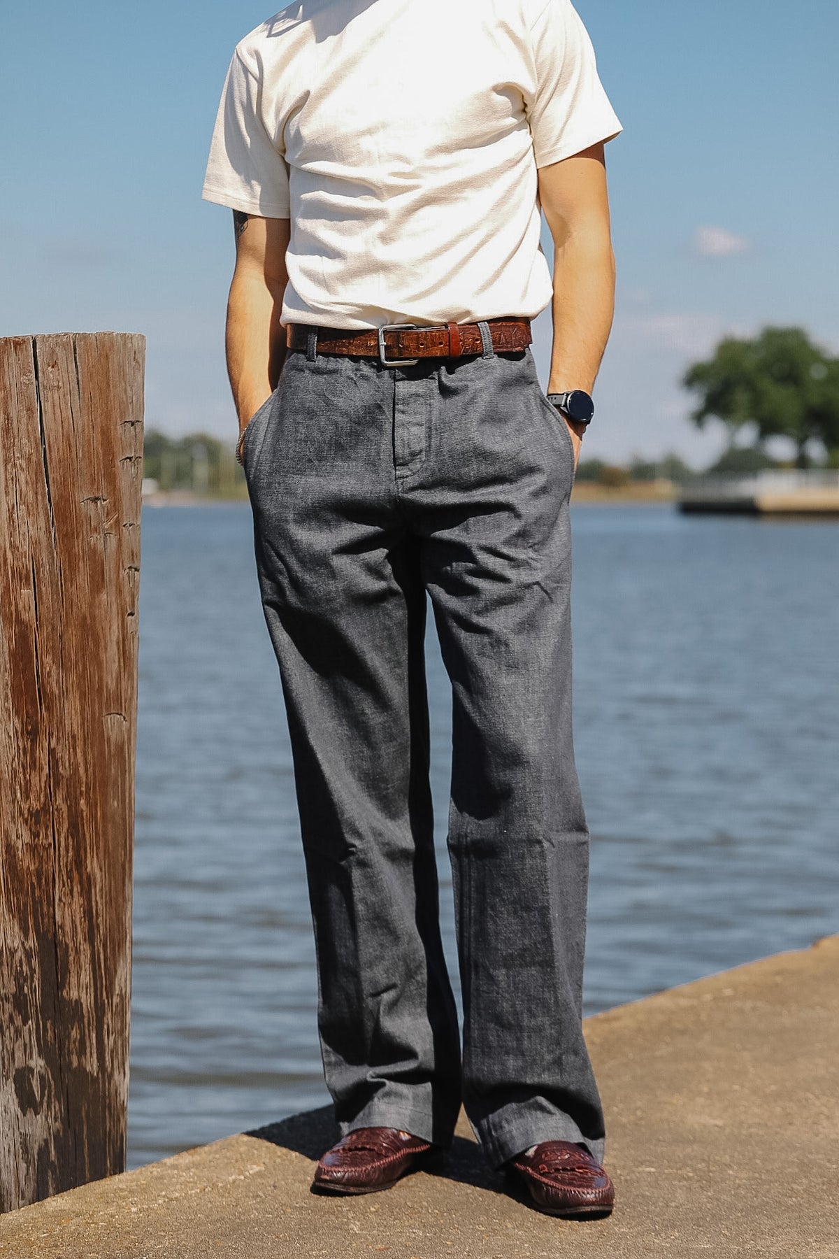 A person standing outdoors by the water wearing loose dark gray trousers, a white short-sleeve shirt, a brown belt, and burgundy loafers.