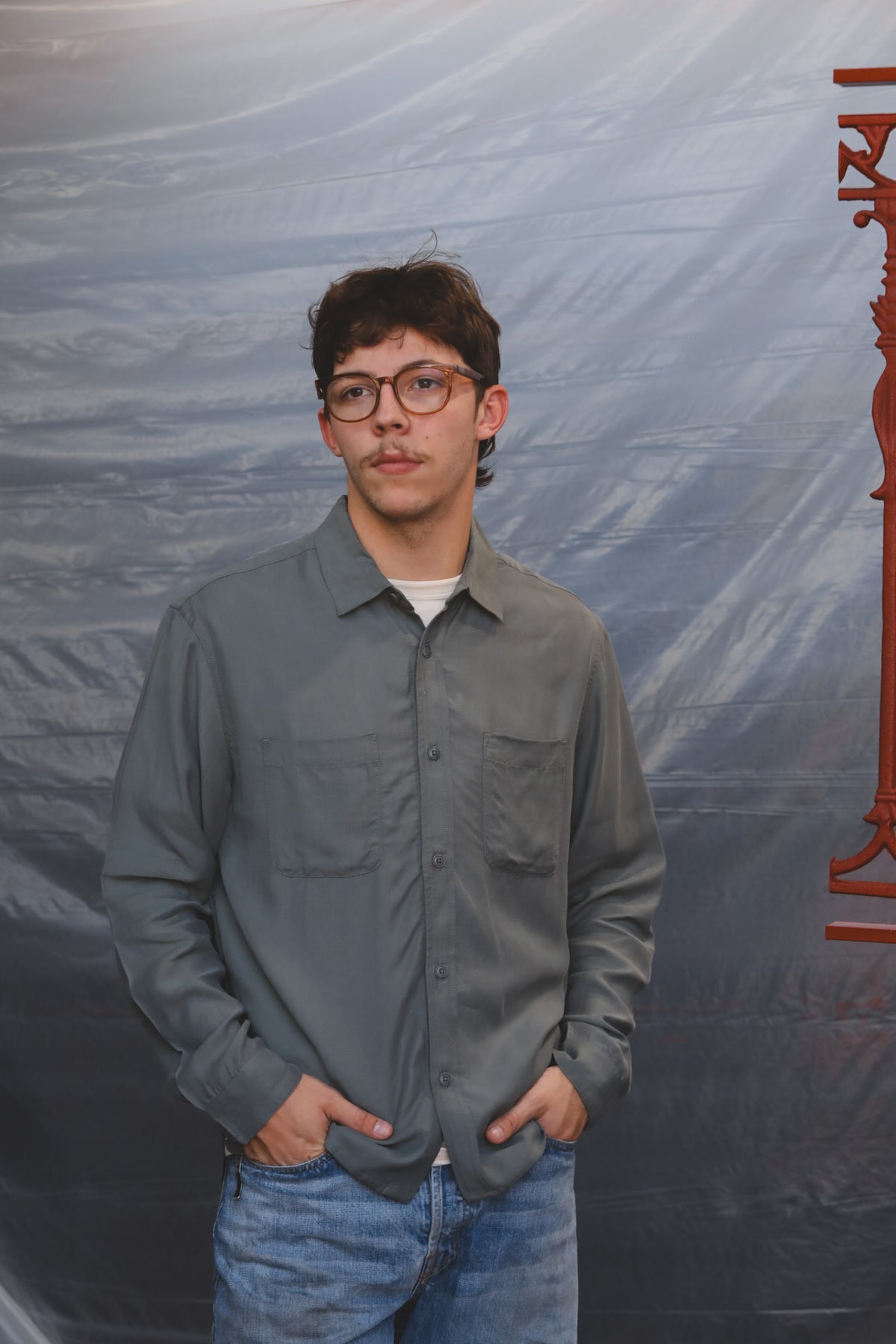 A young man with glasses and a mustache stands wearing a gray button-up shirt and blue jeans, posing in front of a dark gray backdrop with a red graphic on the right.