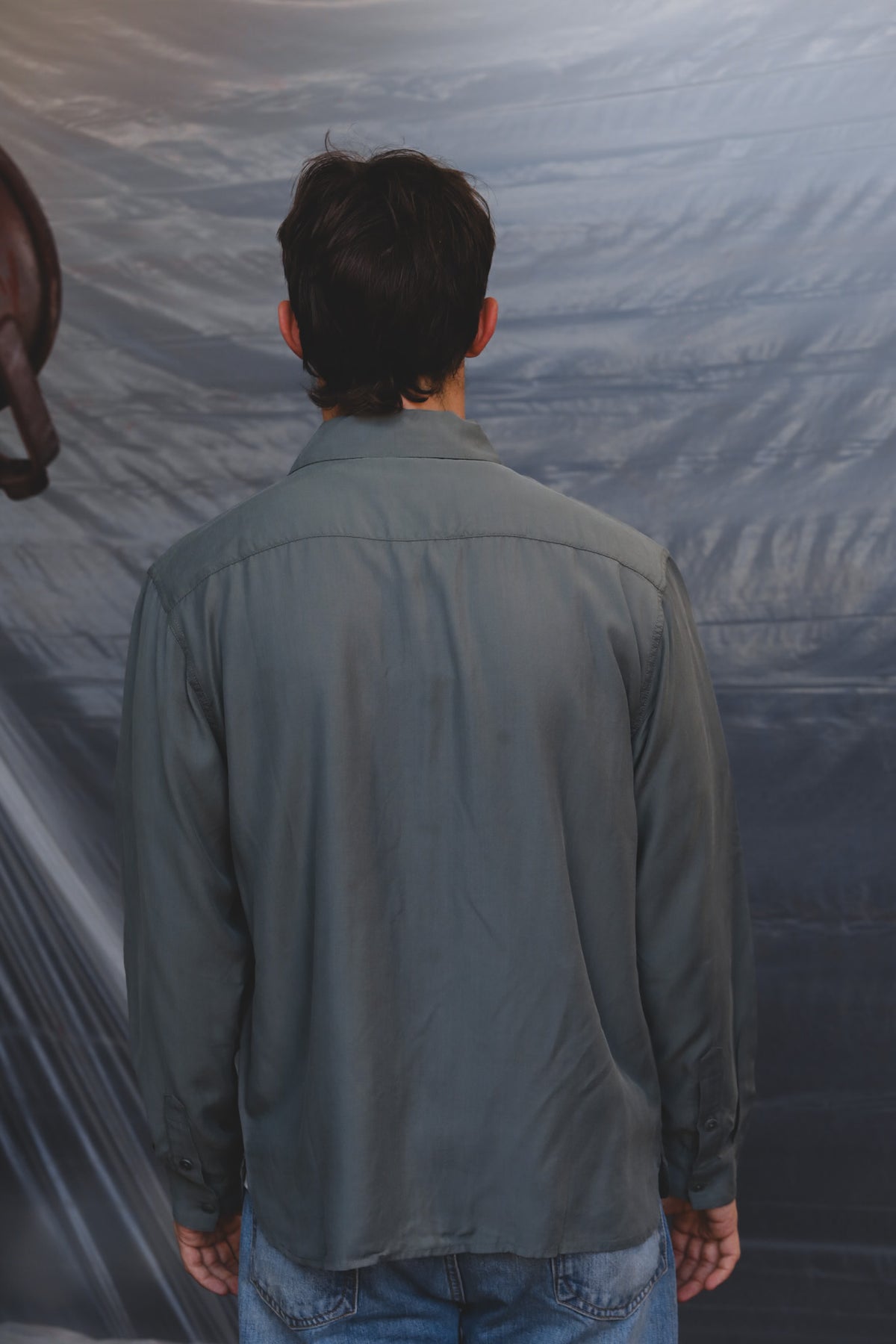 A man viewed from behind wearing a muted gray-green long-sleeve button-up shirt and blue jeans, standing in front of a wrinkled gray backdrop.