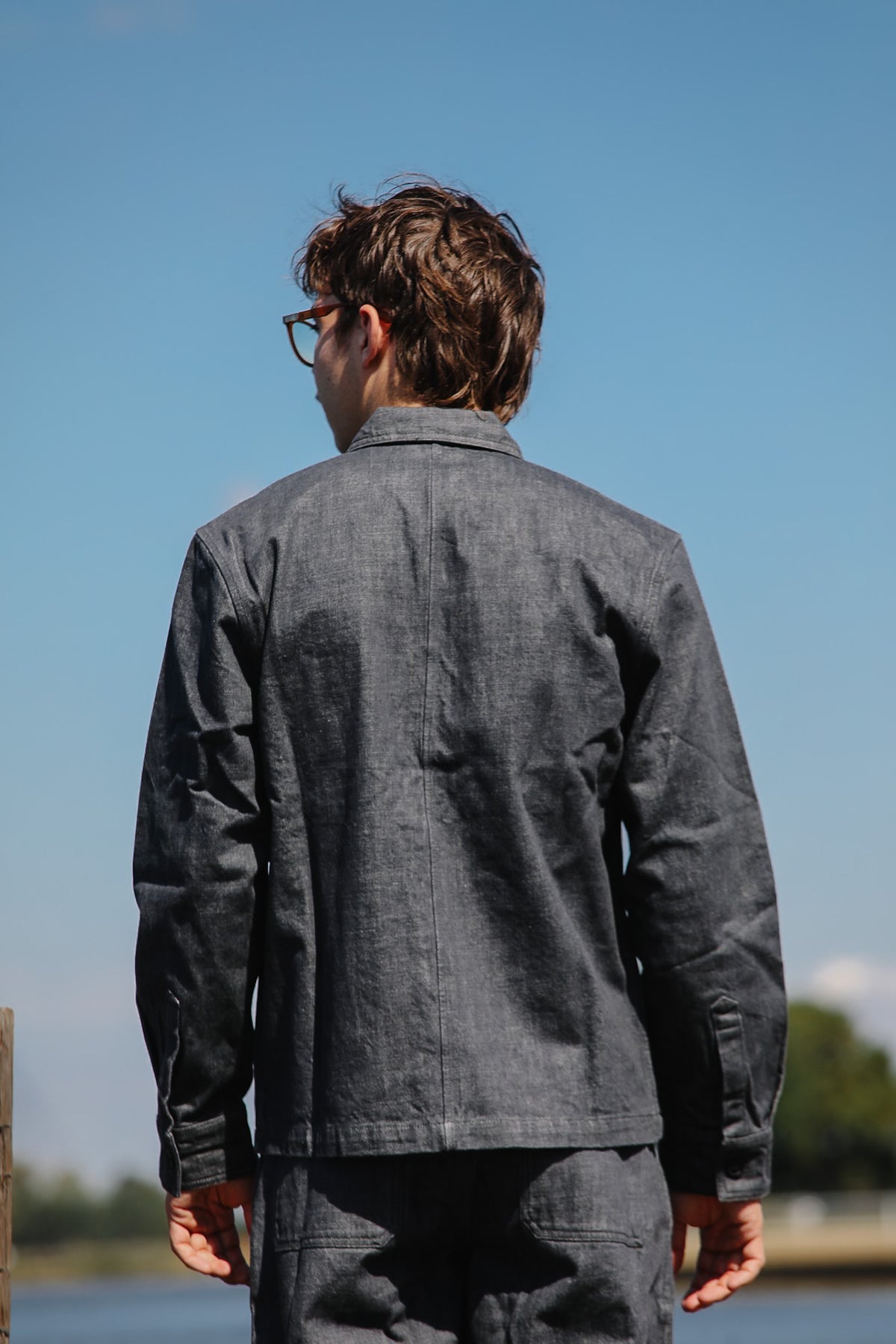 A man viewed from behind wearing a dark gray textured linen jacket and matching trousers, standing outdoors against a clear blue sky.