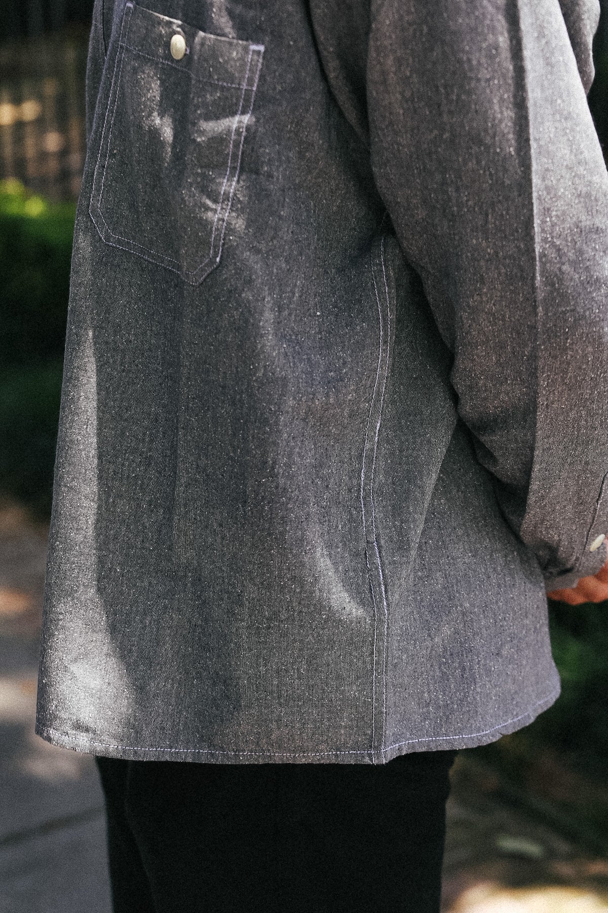 Close-up of a person wearing a dark gray textured overshirt or jacket with a chest pocket, paired with black pants, outdoors.