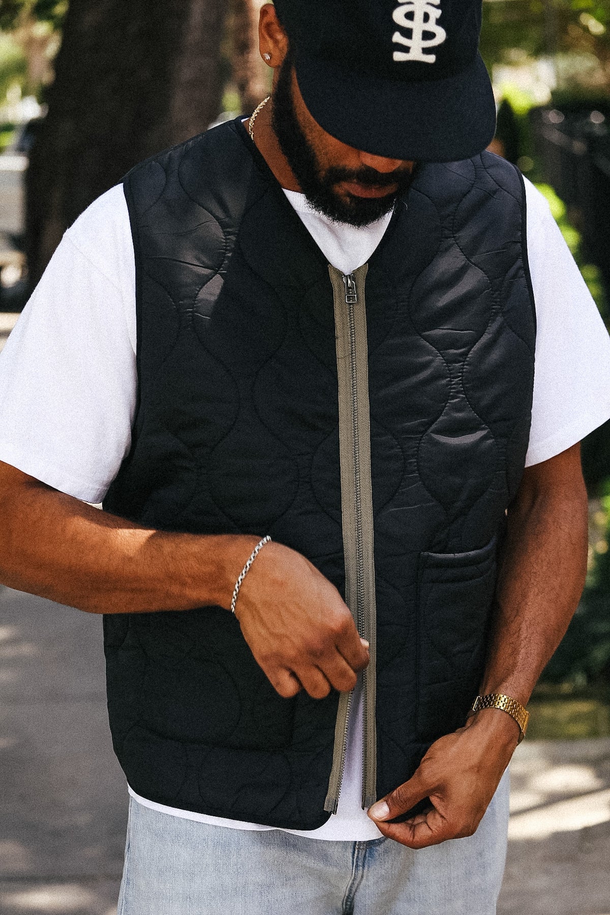 A man wearing a black quilted zip-up puffer vest over a white t-shirt and light blue jeans, standing outdoors in a park-like setting.